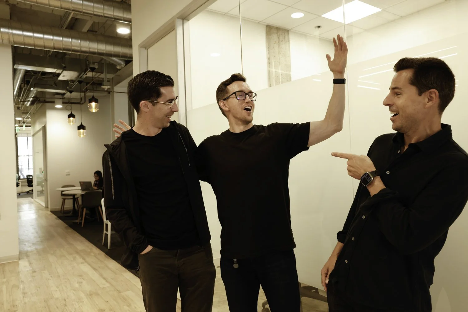 Three men laughing and joking in a modern office hallway, with one pointing at another at Brex Headquarter in San Francisco, CA.