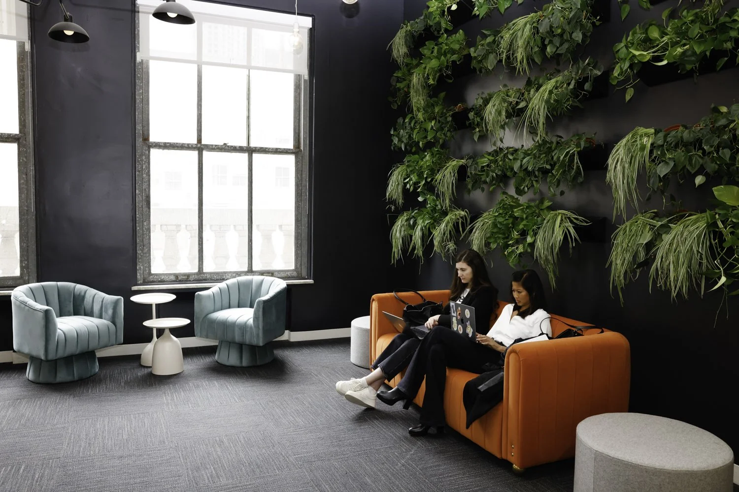 Two women sitting on an orange sofa in a modern lounge area, working on laptops. The room has large windows, two light blue velvet chairs, and a wall with green plants Brex headquarter in San Francisco, CA.