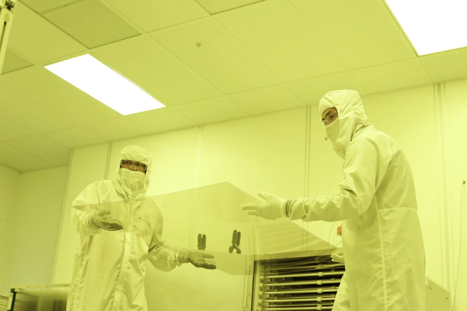 Two scientists carrying a glass plate in bunny suits and goggles under yellow light in San Francisco, CA. 