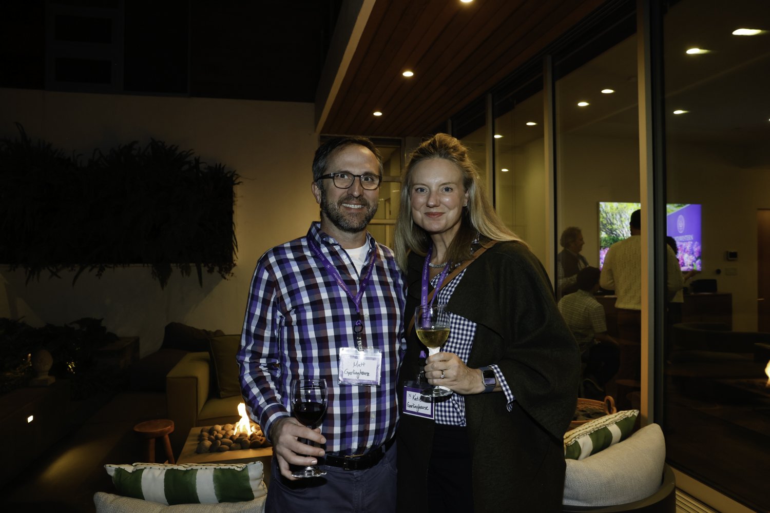 A man and woman smiling at a social event, holding glasses of wine in an indoor setting with modern decor and a fireplace in the background.
