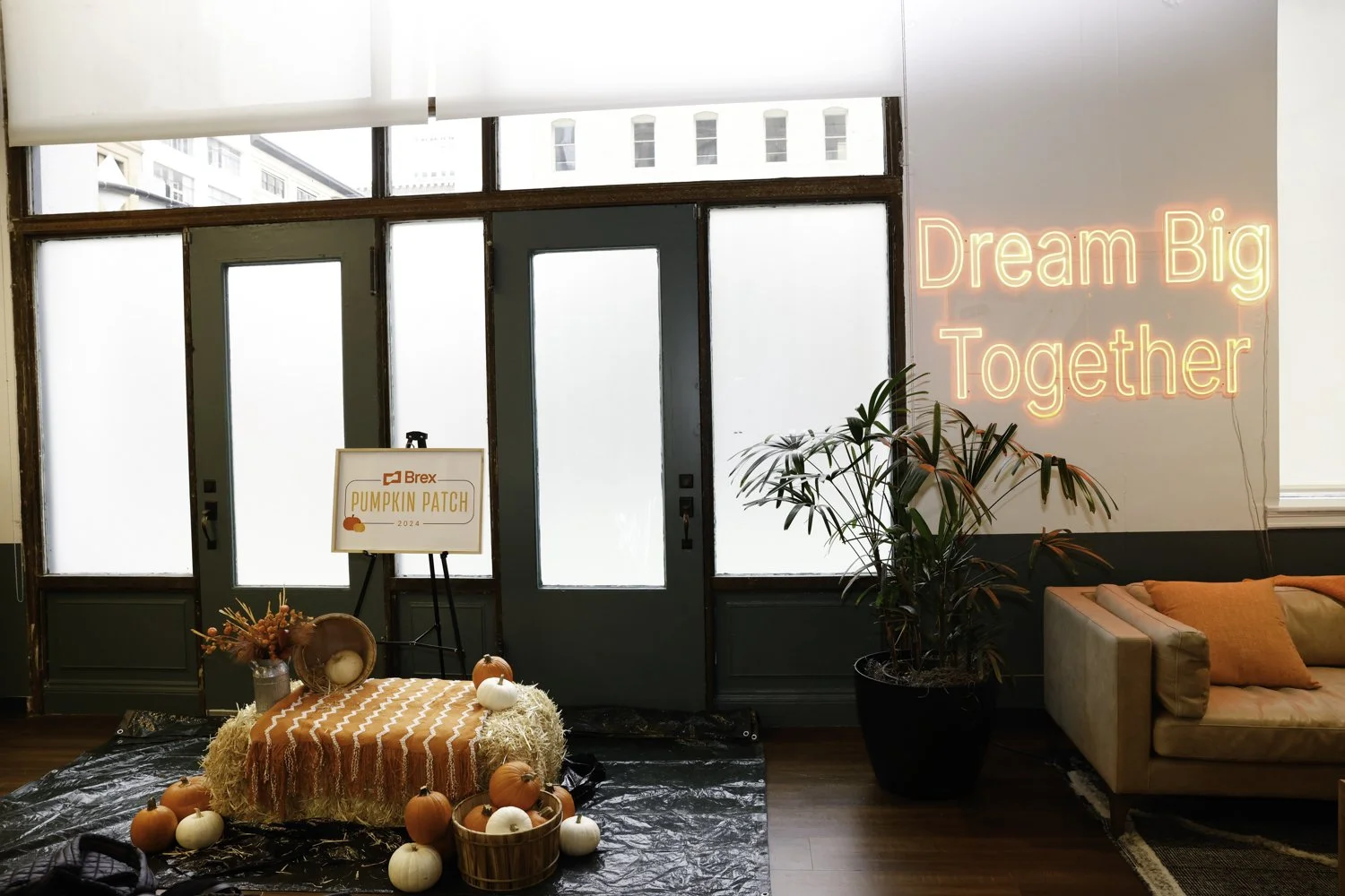 Interior of a room with large windows, decorated for autumn with pumpkins, a hay bale, and flowers on a table. A sign reads 'Brex Pumpkin Patch 2024'. A lime green door is in the background. On the right wall, a neon sign says 'Dream Big Together'. A