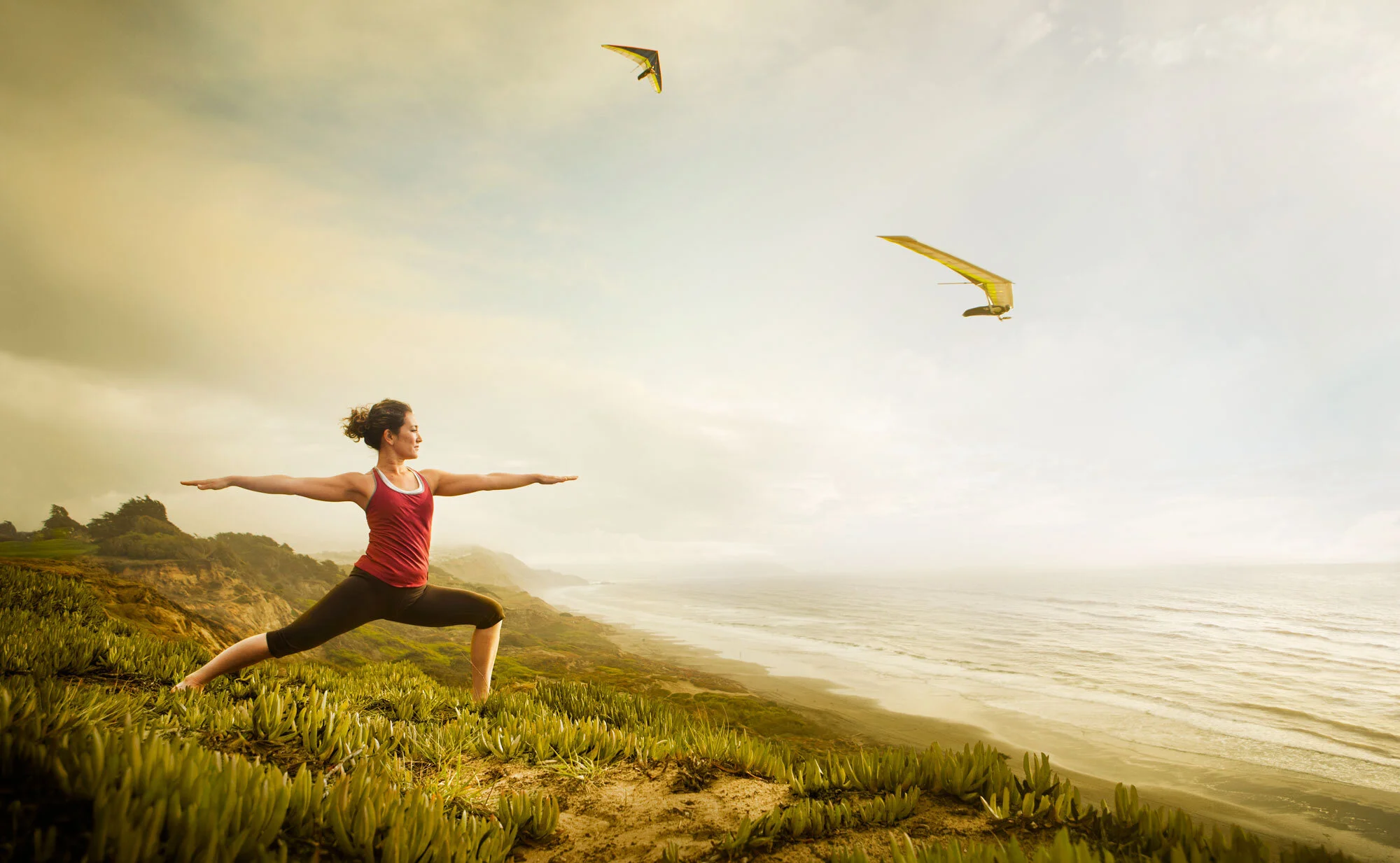Japanese and Spanish woman practicing yoga on a coastal cliff at Ocean Beach with two yellow hang gliders flying above her in San Francisco, CA.