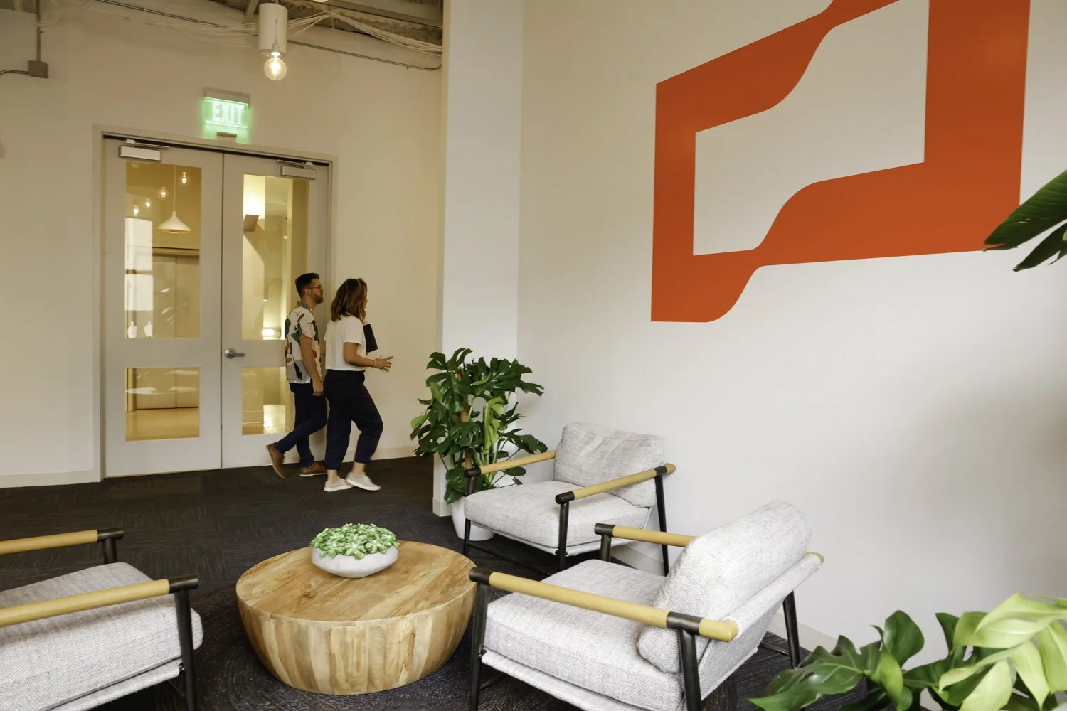 Two people walking into a modern office lobby with white walls, potted plants, and contemporary furniture, including two gray armchairs and a round wooden coffee table with a plant on it. A large red and white abstract wall art is on the wall.