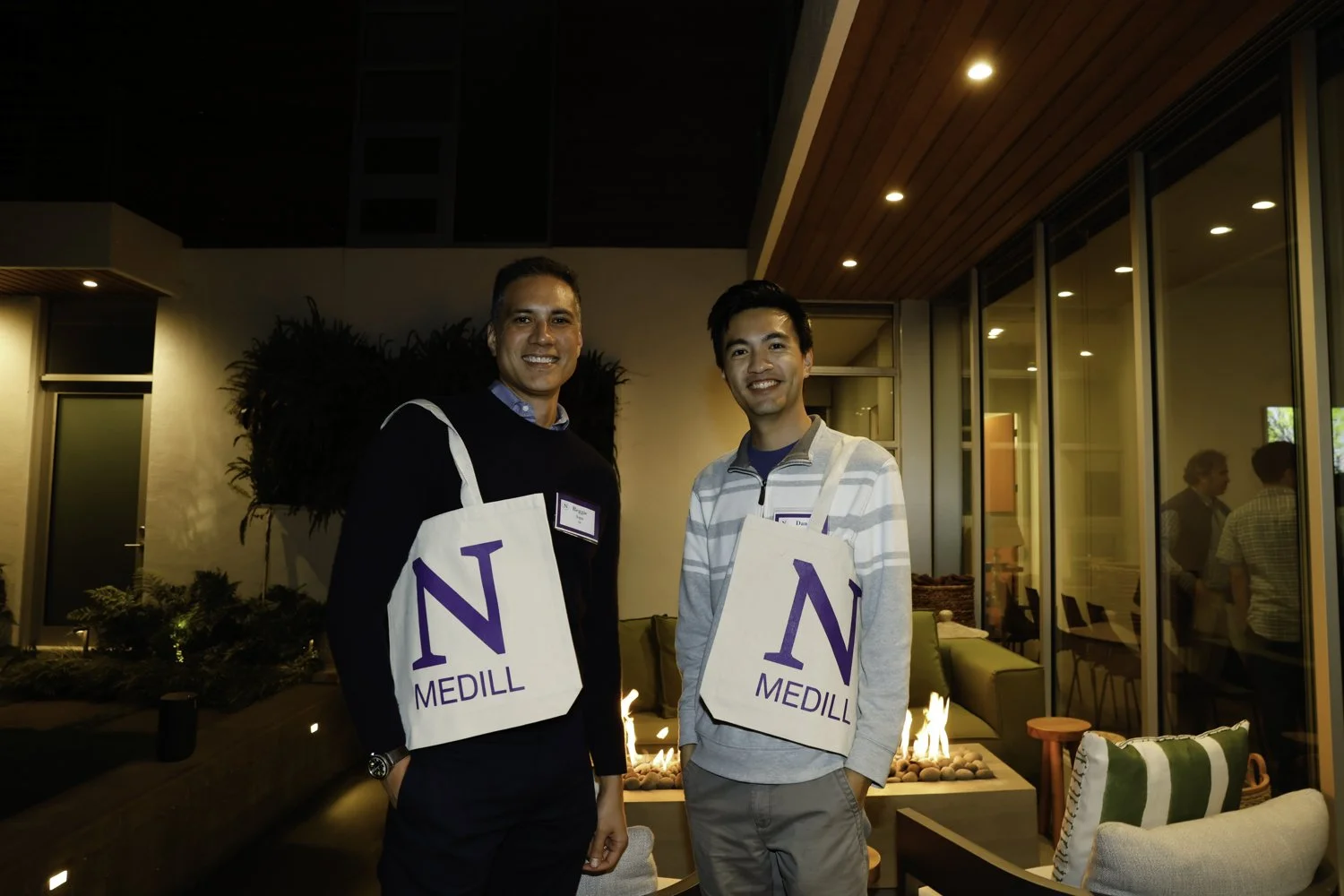 Two men smiling at a social event indoors, wearing tote bags with the letter 'N' and the word 'Medill', in a warmly lit room with glass walls and chairs at Northwestern fundraiser in San Francisco, CA.