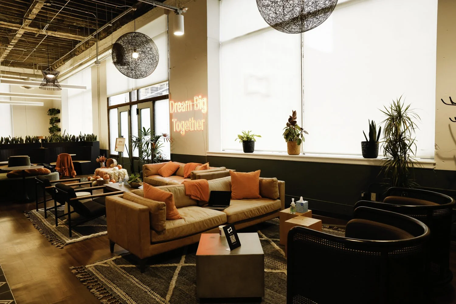 Cozy modern lounge area with beige couches, orange pillows and throws, black armchairs, indoor plants, large window, neon sign that reads "Dream Big Together", wooden tables, and decorative lighting Brex headquarter in San Francisco, CA.
