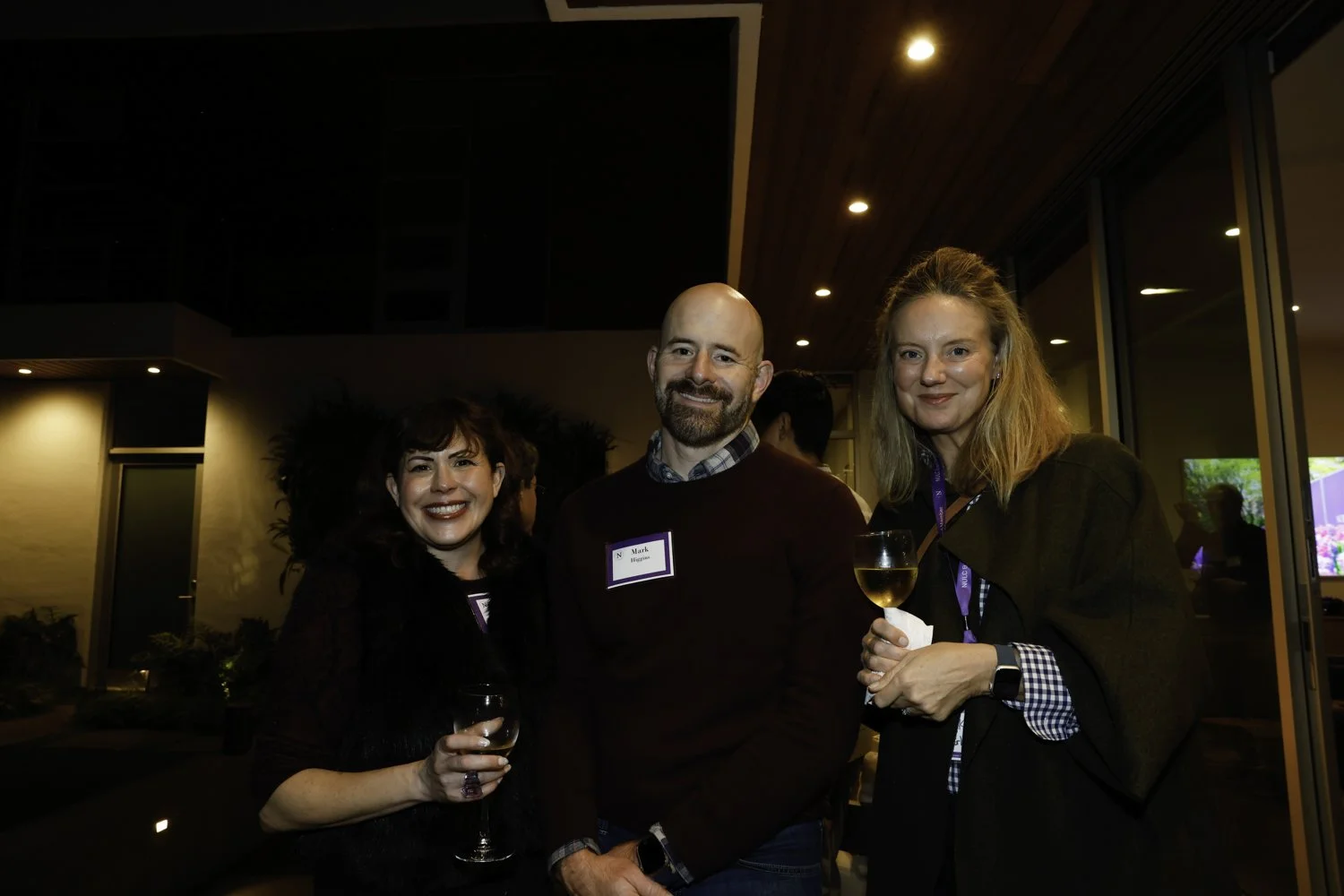 Three people at an indoor social event, smiling, with two women holding glasses of white wine and a man with a name tag that says 'Mark' at Northwestern fundraiser in San Francisco, CA.