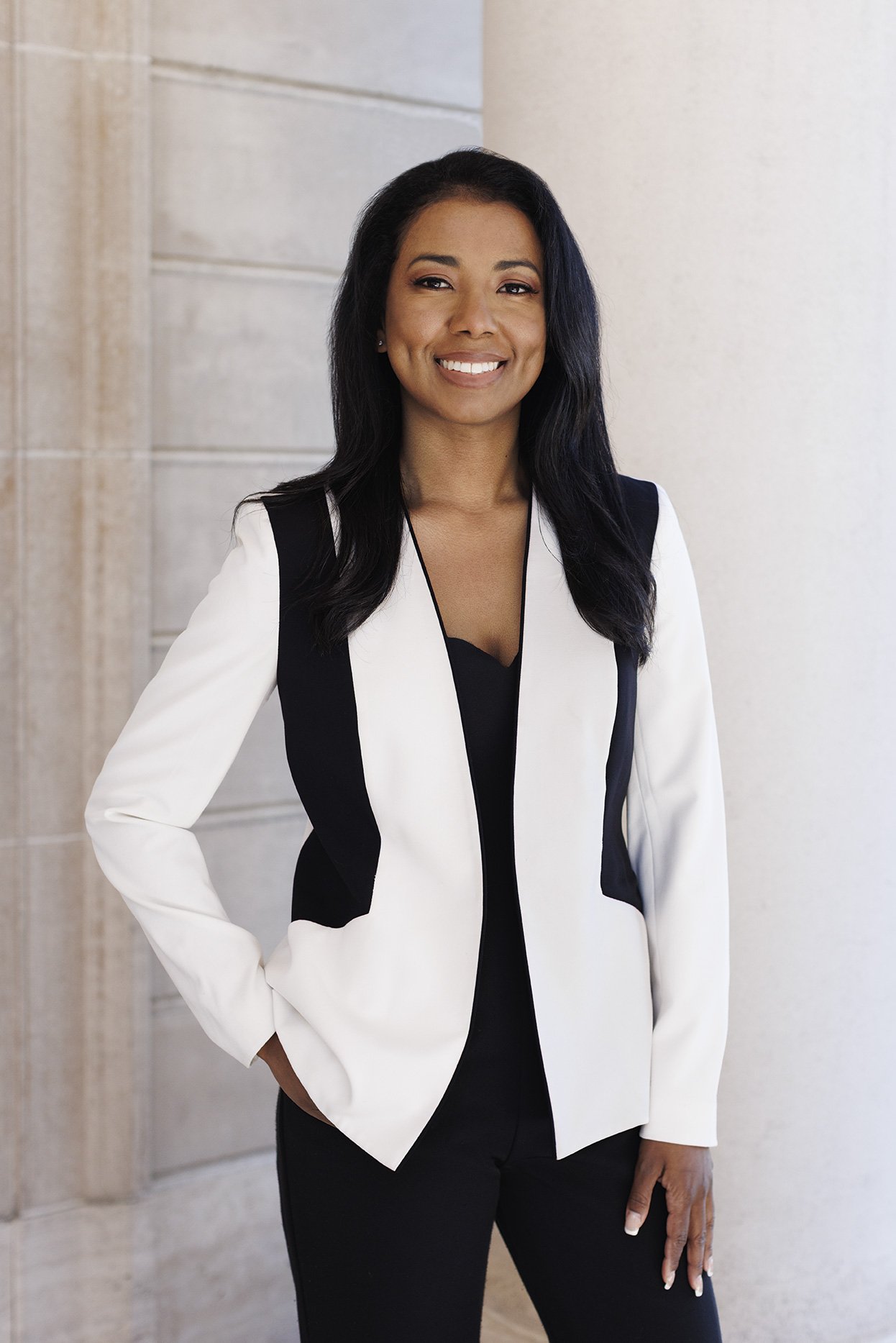 Young woman dressed in a black shirt and white and black blazer and black pants, with one hand in her pocket, against a soft white and grey stonewall in the Presidio, in San Francisco, CA.