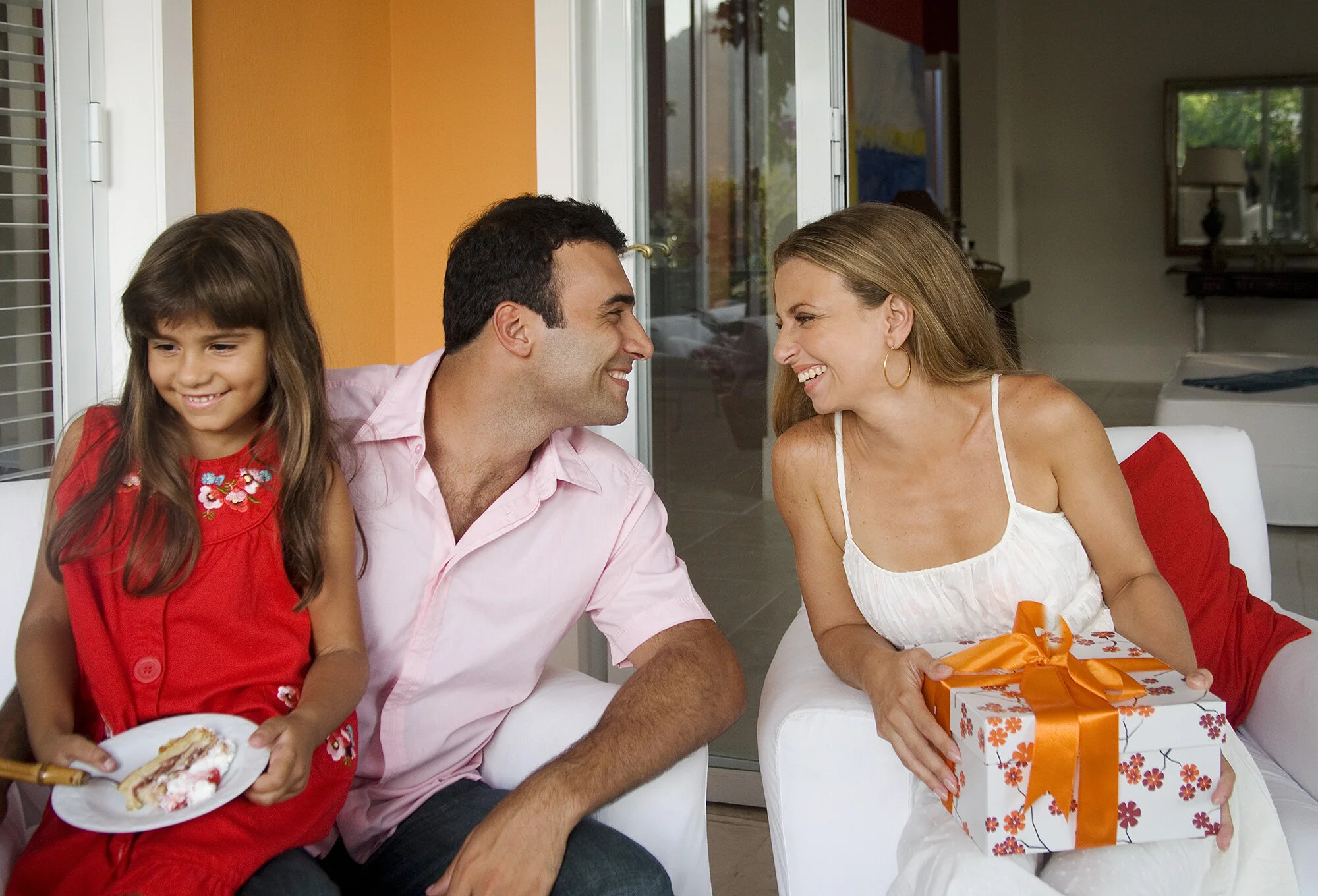 A family celebrating a birthday, featuring a young girl in a red dress with floral embroidery holding a plate with a cake, a man in a pink shirt, and a woman in a white dress with a gift wrapped in floral paper and orange ribbon, all smiling and sitt