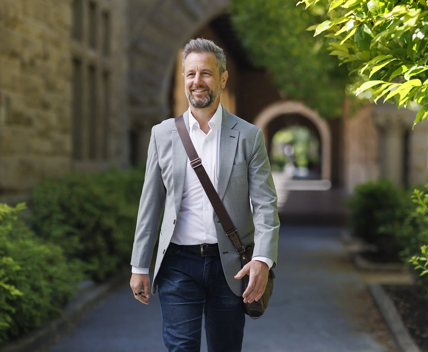 A middle-aged man in a gray blazer and white shirt walking outdoors along a path with greenery and trees with a brick arch behind him at Stanford University Campus at Stanford, CA.