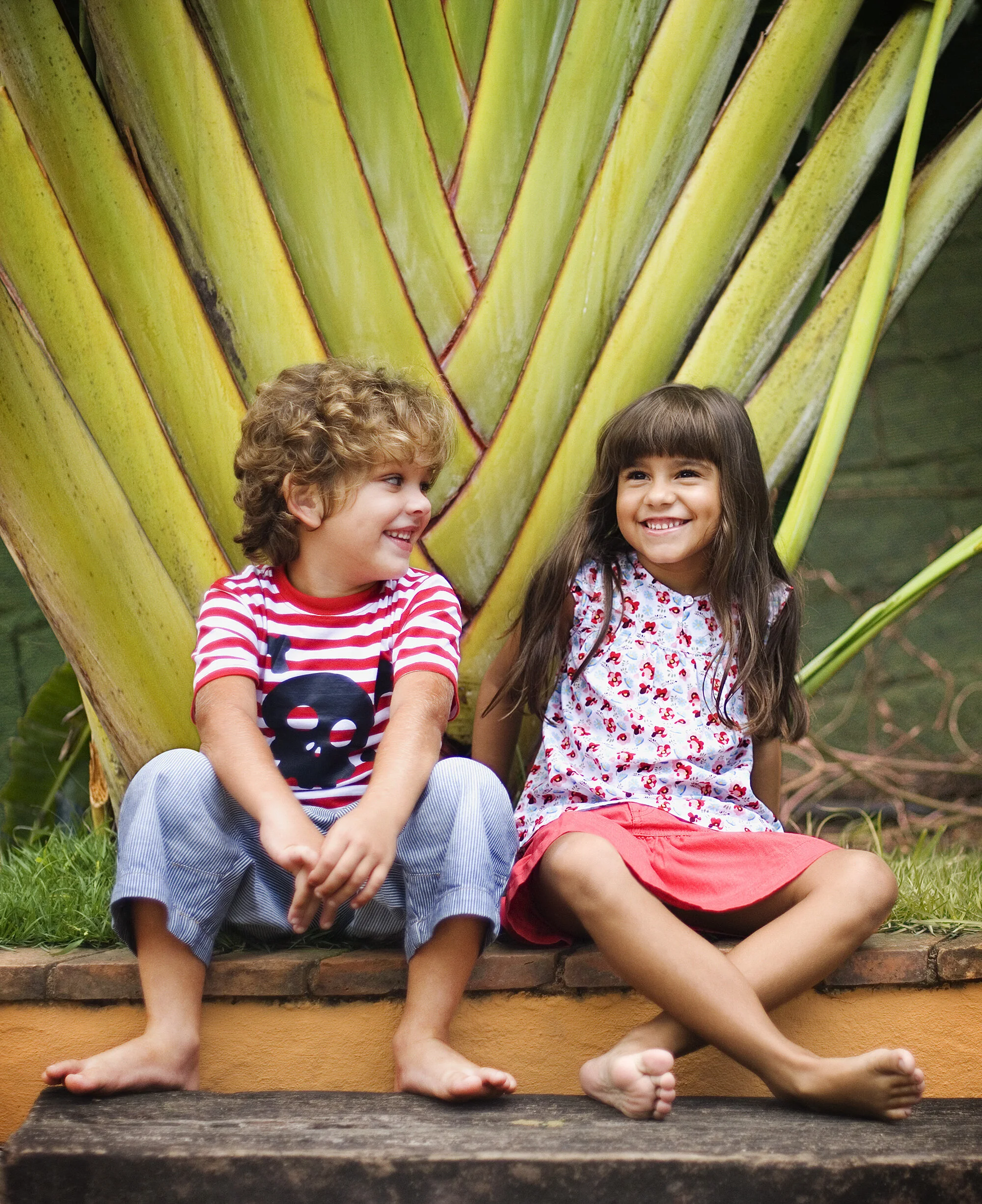 Two Brazilian children, a boy with curly blonde hair and a girl with long brown hair, sitting barefoot on a wooden surface in front of a large banana plant, smiling and looking at each other in Rio de Janeiro, Brazil.
