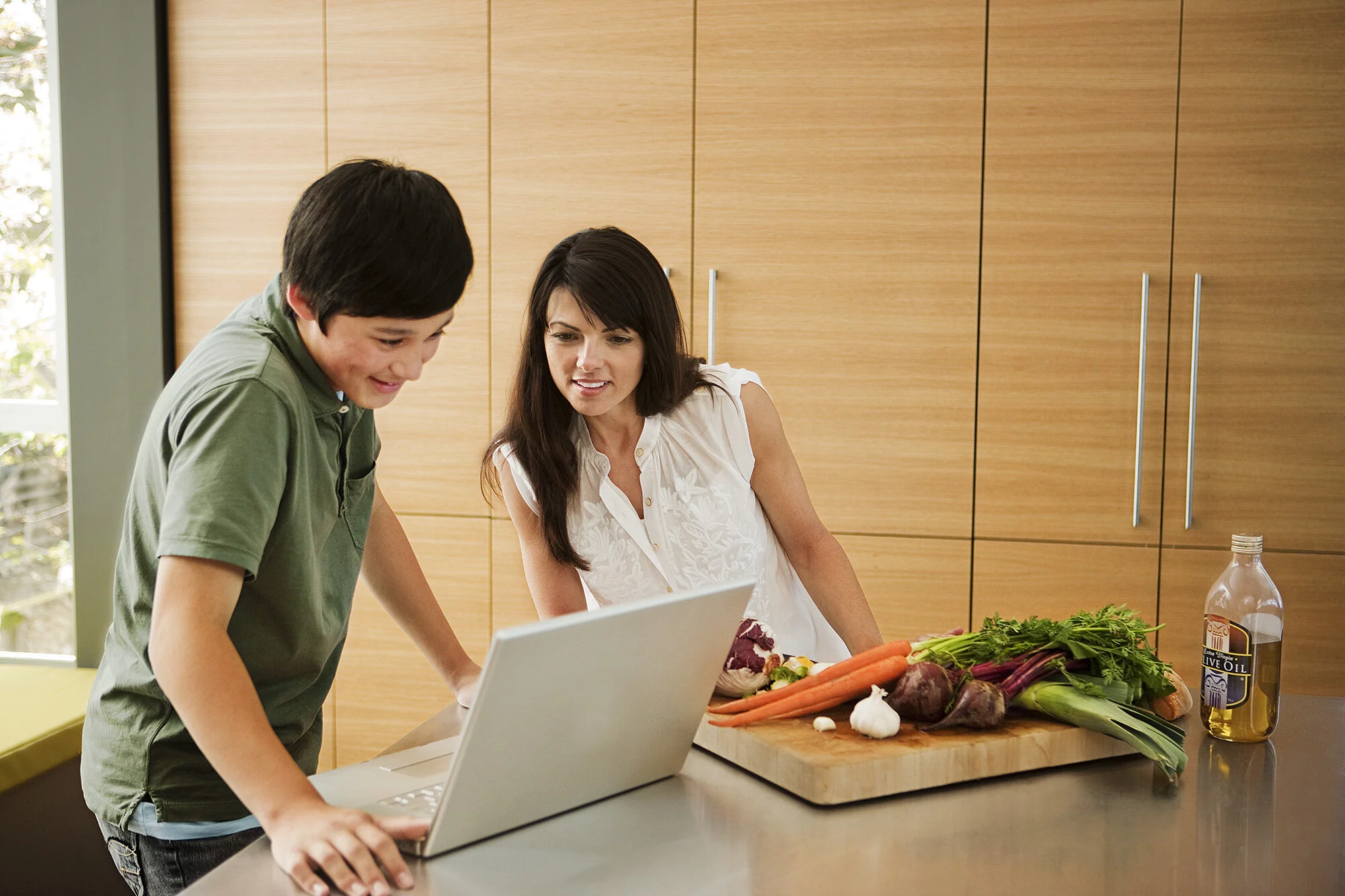 A mother and a son looking at a laptop on a kitchen counter with fresh vegetables and a bottle of olive oil. The mother is wearing a green shirt, and the son is wearing a white sleeveless blouse in San Francisco, CA.