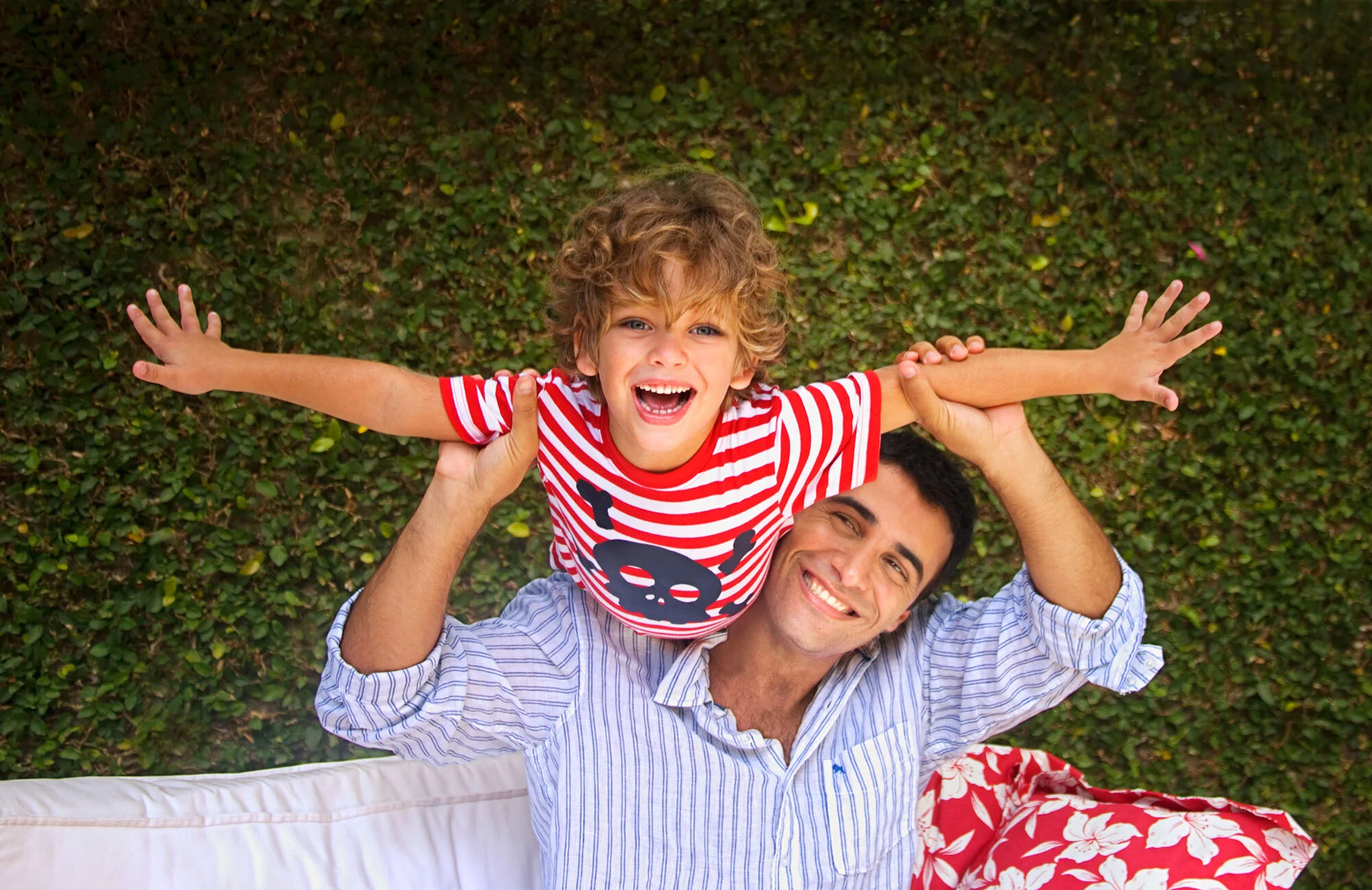 A man lying on a white lounge chair on green grass, holding a smiling young boy above his head with outstretched arms. The boy is wearing a red and white striped shirt with a black skull graphic and has curly blonde hair and a big smile. The man is w