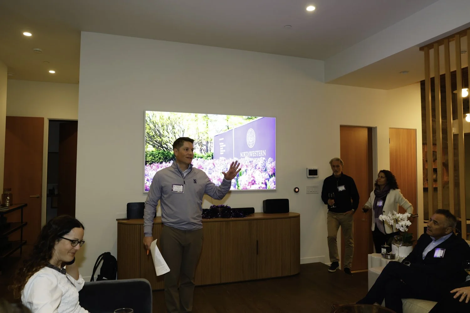 A man in a gray shirt giving a presentation to a small group in a modern room with a large screen displaying 'NORTHWESTERN' and a purple floral background. Several people are seated and standing, listening and drinking wine at Northwestern fundraiser