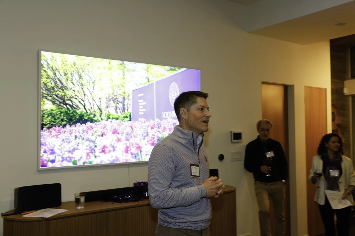 Man giving a speech at a conference with a large screen behind him displaying a garden scene and a purple sign. Two individuals are standing nearby, one holding a glass of wine and the other holding papers at Northwestern fundraiser in San Francisco,