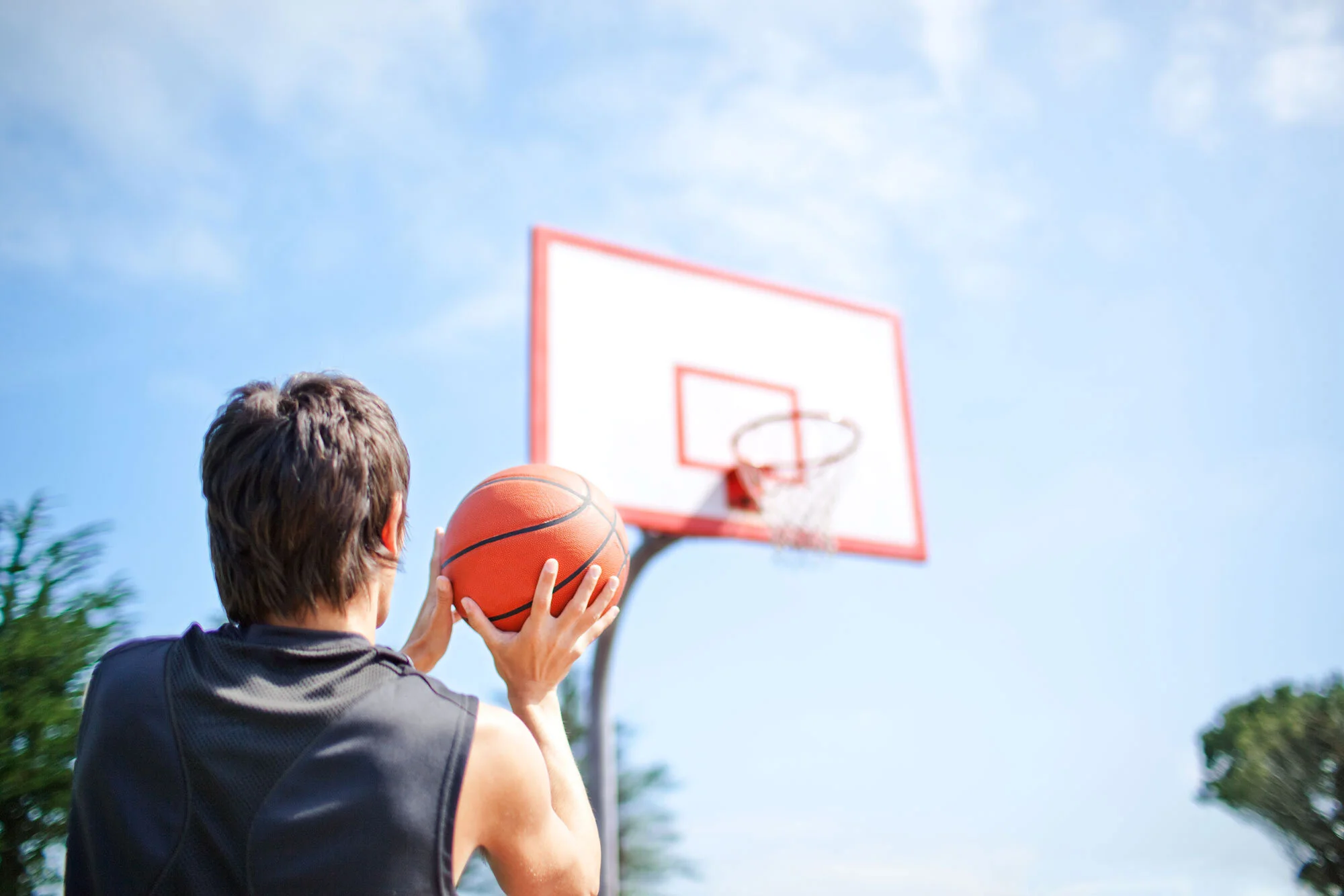 A person with short brown hair holding a basketball, preparing to shoot towards a basketball hoop outdoors on a sunny day with a blue sky and some clouds n San Francisco, CA.