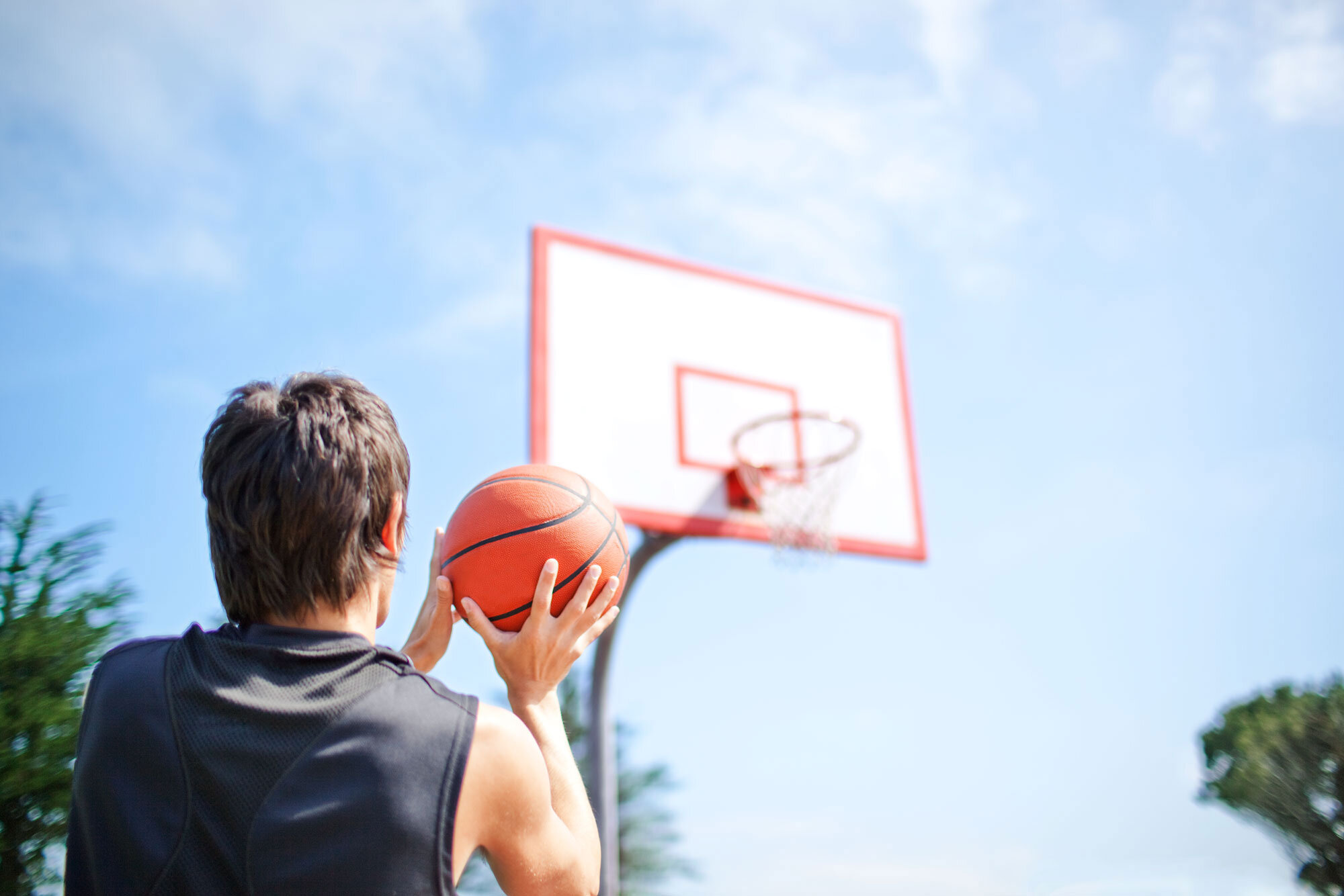 A young Argentinian man with short brown hair holding a basketball, preparing to shoot towards a basketball hoop outdoors on a sunny day with a blue sky and some clouds n San Francisco, CA.