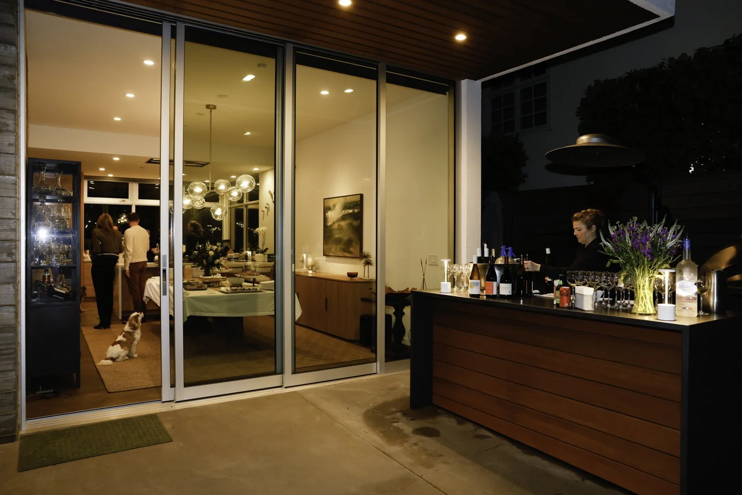 View of a party or gathering inside a modern house seen through glass sliding doors, with a woman preparing drinks at an outdoor bar, and people inside near a dining table with a dog sitting on a mat Brex headquarter in San Francisco, CA.