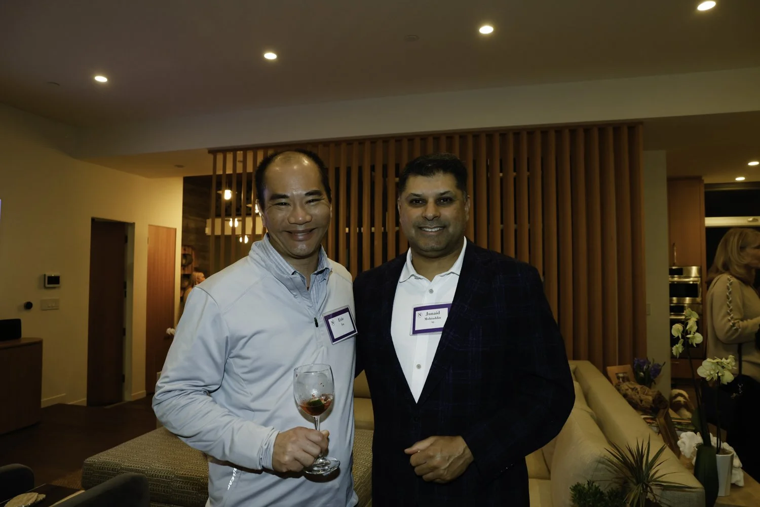 Two men standing together at a social event, smiling. The man on the left is holding a glass of red wine and wearing a light blue shirt. The man on the right is dressed in a dark blazer over a white shirt. They are in a cozy, warmly lit room with woo