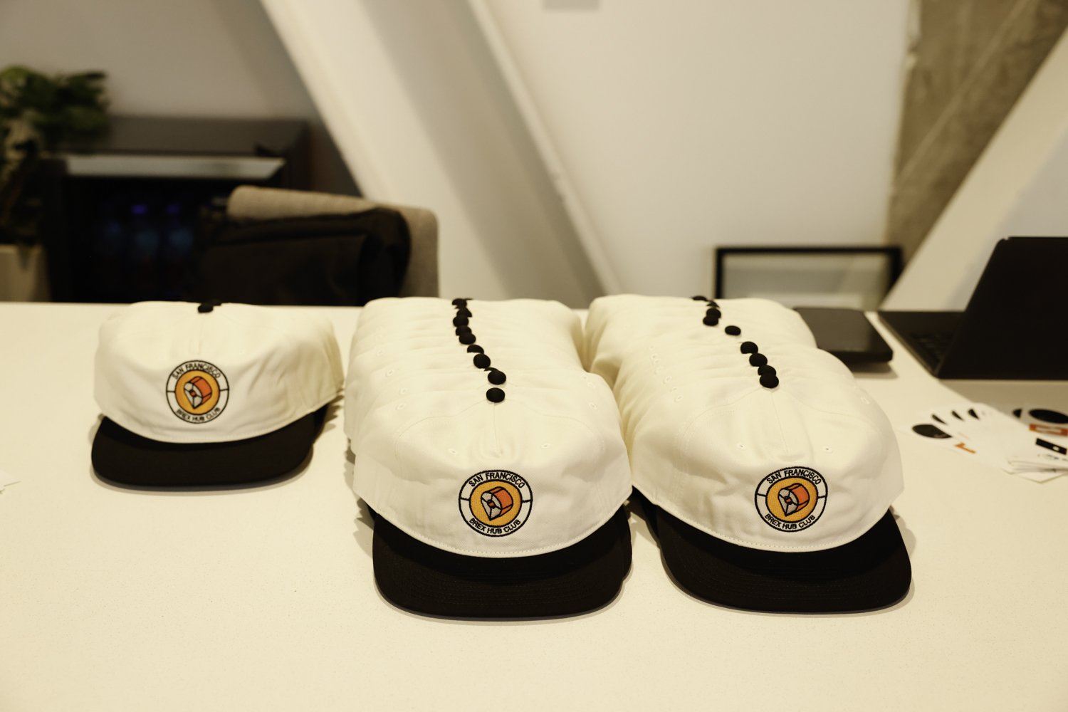 Three white caps with black brims and black buttons, featuring a San Francisco Bay Hub Club logo, are placed on a white surface, with a snack and laptop in the background Brex headquarter in San Francisco, CA.