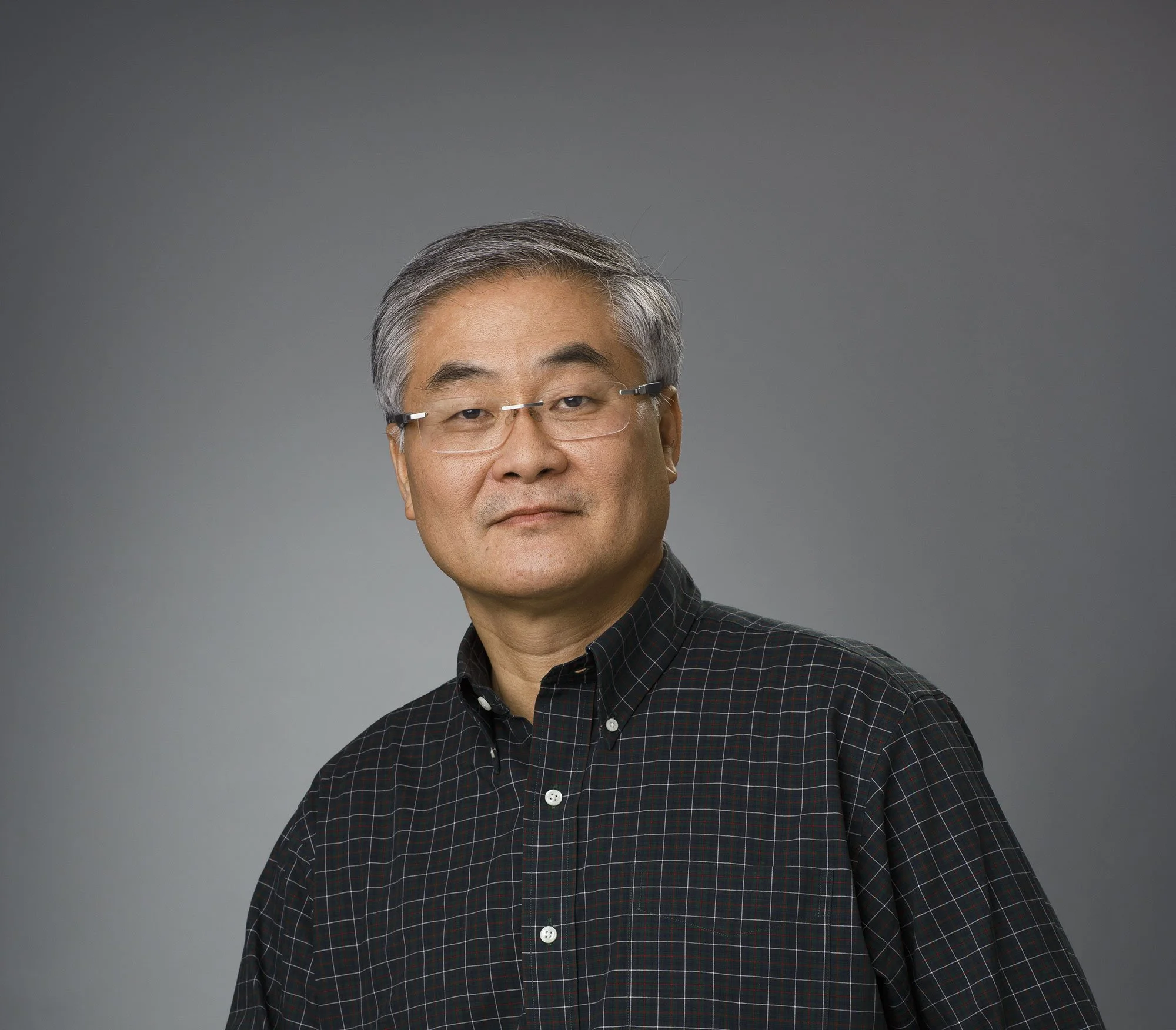 Portrait of a middle-aged Asian man with short gray hair and glasses, wearing a black checkered shirt, against a plain gray background in San Francisco, CA.