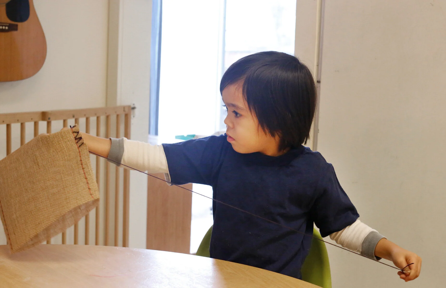 A young boy with dark hair, wearing a navy blue shirt and a long-sleeve white shirt underneath, is sitting at a table. He is holding a string with his left hand attached to a woven bag he is sowing at the The Scandinavian School in San Francisco, CA.