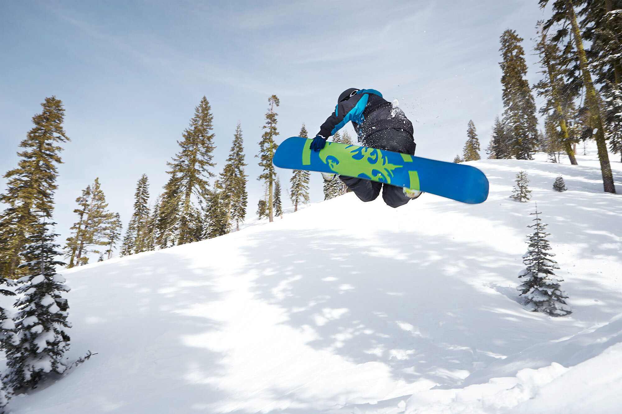 A skier soaring through the air on a snowy mountain slope, surrounded by pine trees under a clear blue sky at Sugar Bowl ski resort, Lake Tahoe, CA.