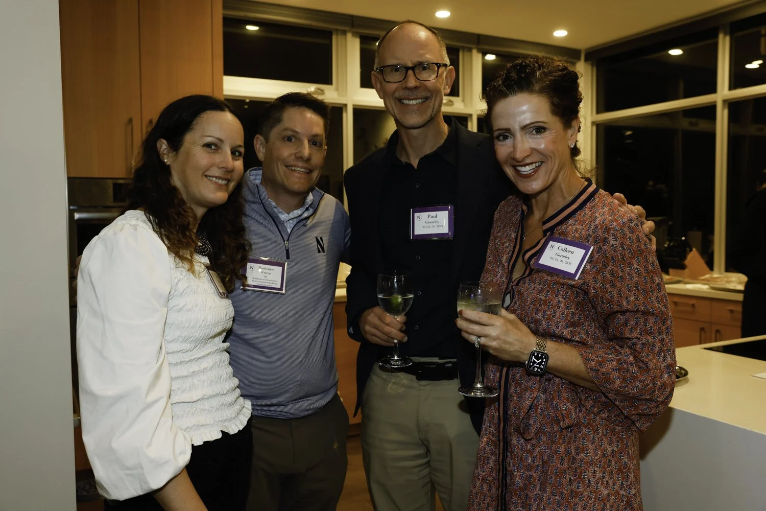 Four people at a social gathering, smiling and holding drinks in a kitchen with wooden cabinets and large windows in the background Brex headquarter in San Francisco, CA.