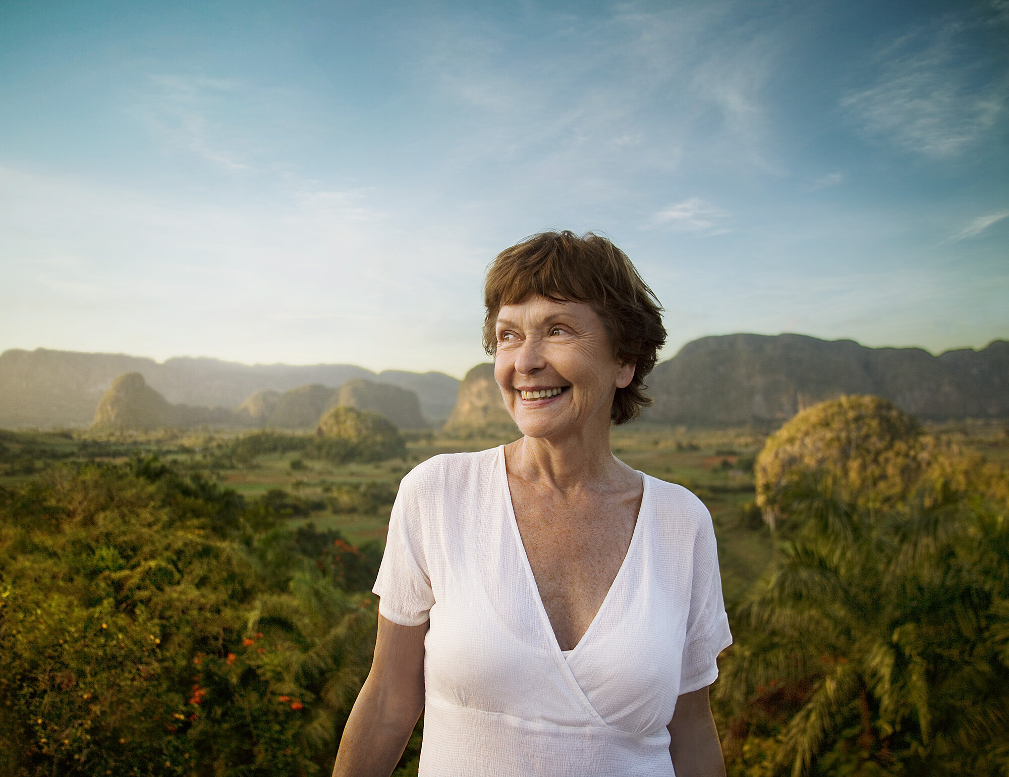 A middle-aged smiling white woman in a white V-neck white shirt standing outdoors with a lush landscape of trees, mountains, and a blue sky in the background in Viñales, Cuba.