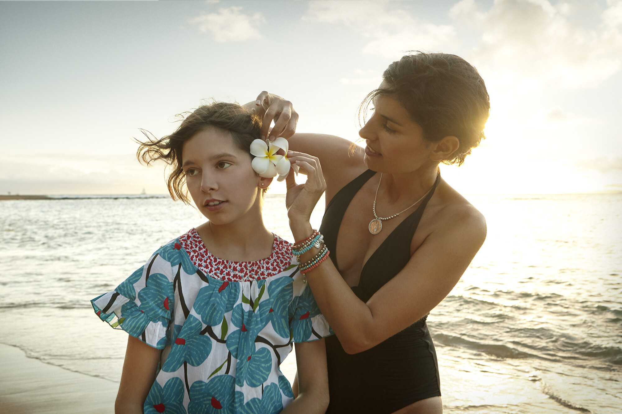 A mother is placing a white flower in her daughter's hair on a beach at sunset with backlit light in Maui, Hawai'i.