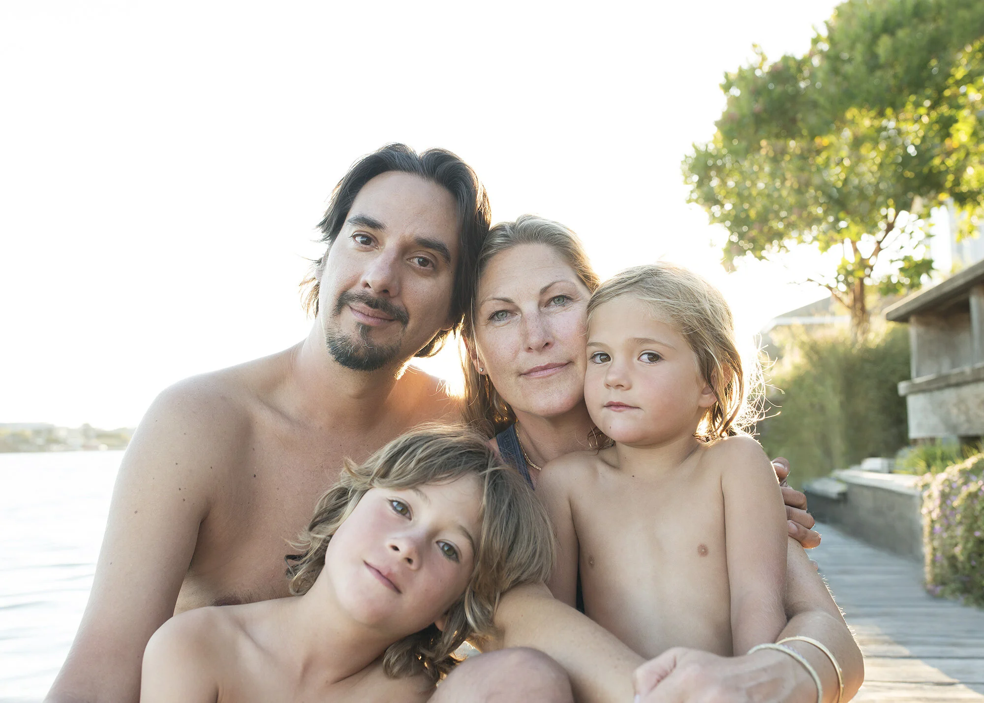 A family of four, including a man, a woman, and two children, sitting together outdoors near a body of water during sunset with bright light in the background at Stinson beach, CA.