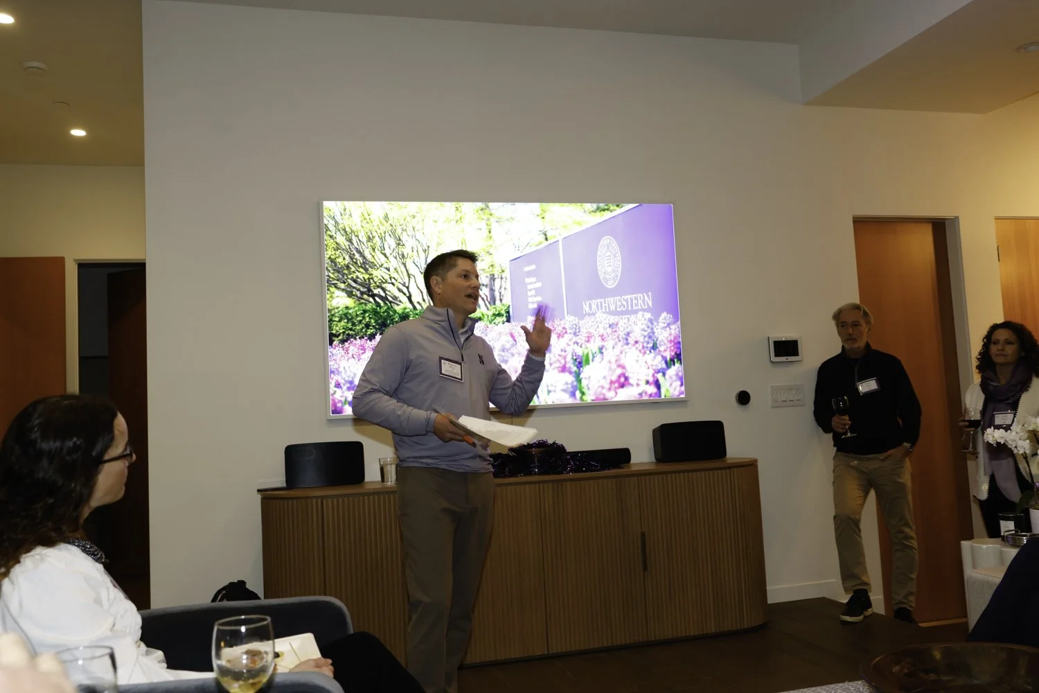 A man giving a presentation in a room with a Northwestern University screen, surrounded by three seated and standing attendees at Northwestern fundraiser in San Francisco, CA.