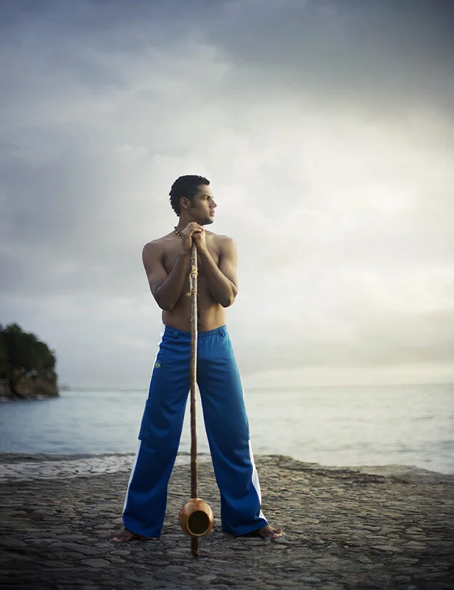 A shirtless Black Brazilian young man with curly hair and blue sweat pants holding a Berimbau, a Brazilian instrument, vertically in front of him, looking to his left with a calm expression, with the ocean and a cloudy sky in the background in Salvad