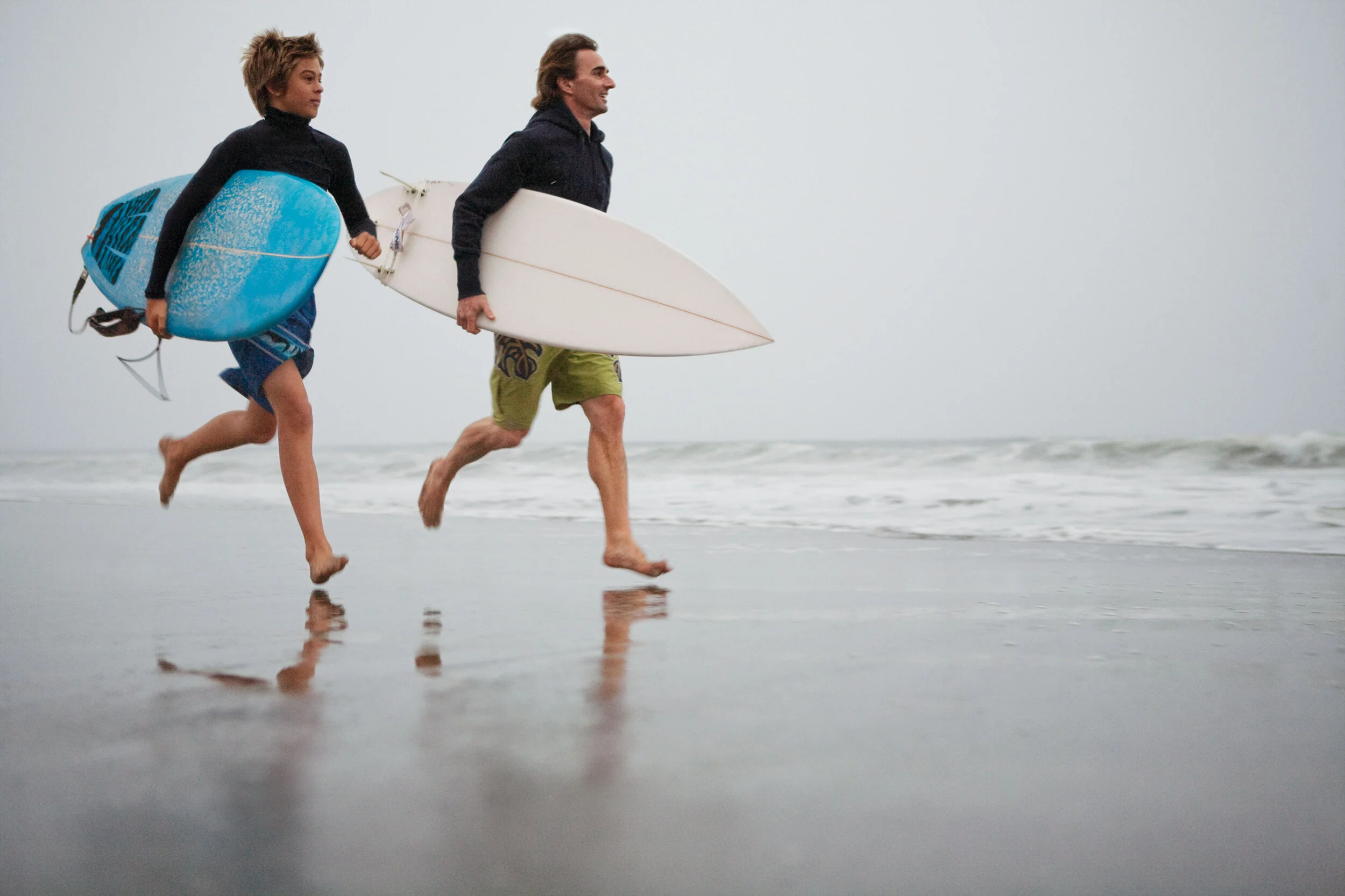 A father and son running along Ocean Beach with surfboards, the boy holding a blue one and the man holding a white one, with the ocean in the background on a cloudy day in San Francisco, CA.