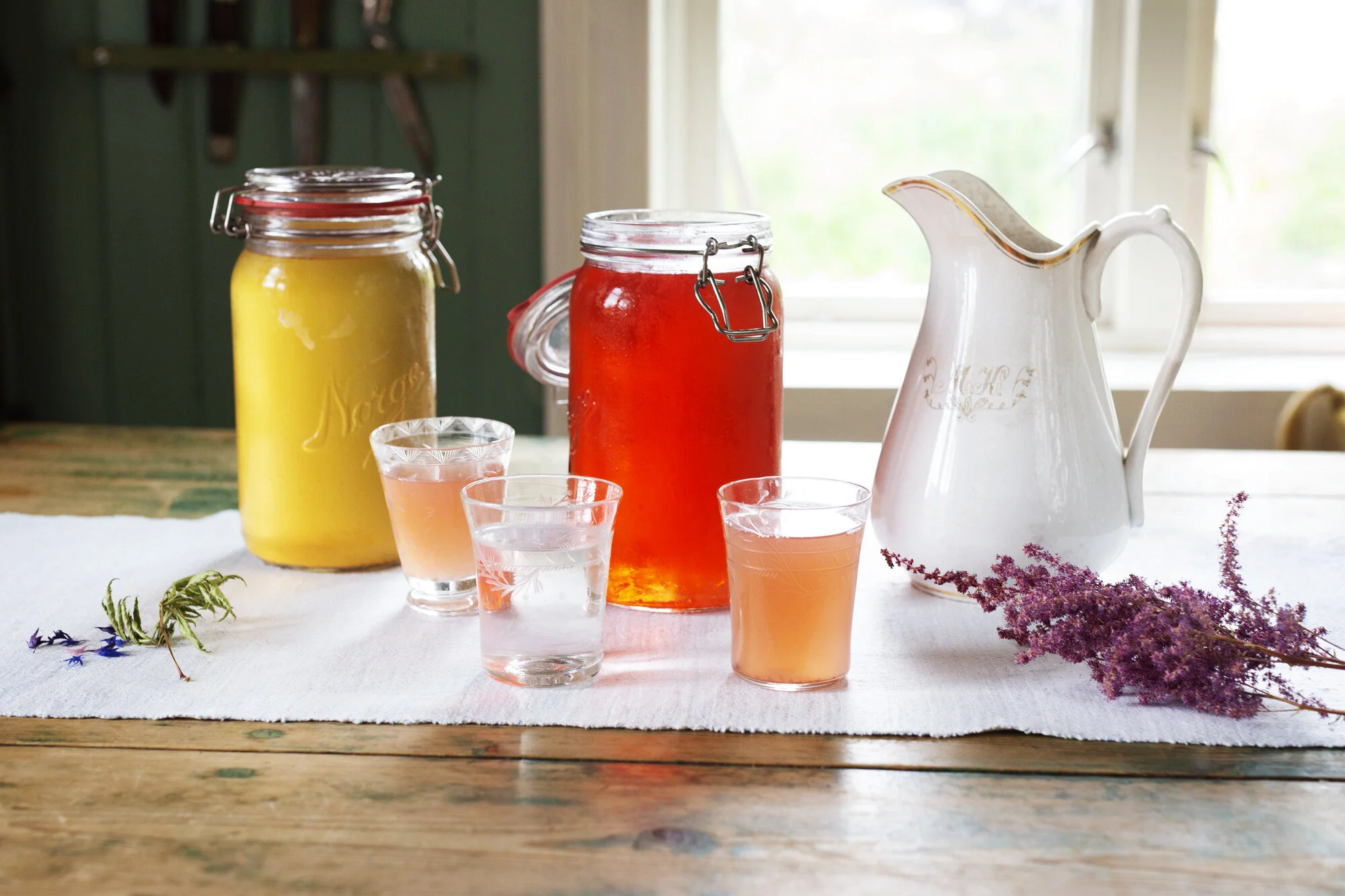 Two large Mason jars filled with lemonade and strawberry juice on a wooden table with three glasses of pinkish juice in a farmhouse. A white ceramic pitcher with gold trim is beside the jars, and a bunch of purple flowers is laid on the table. window