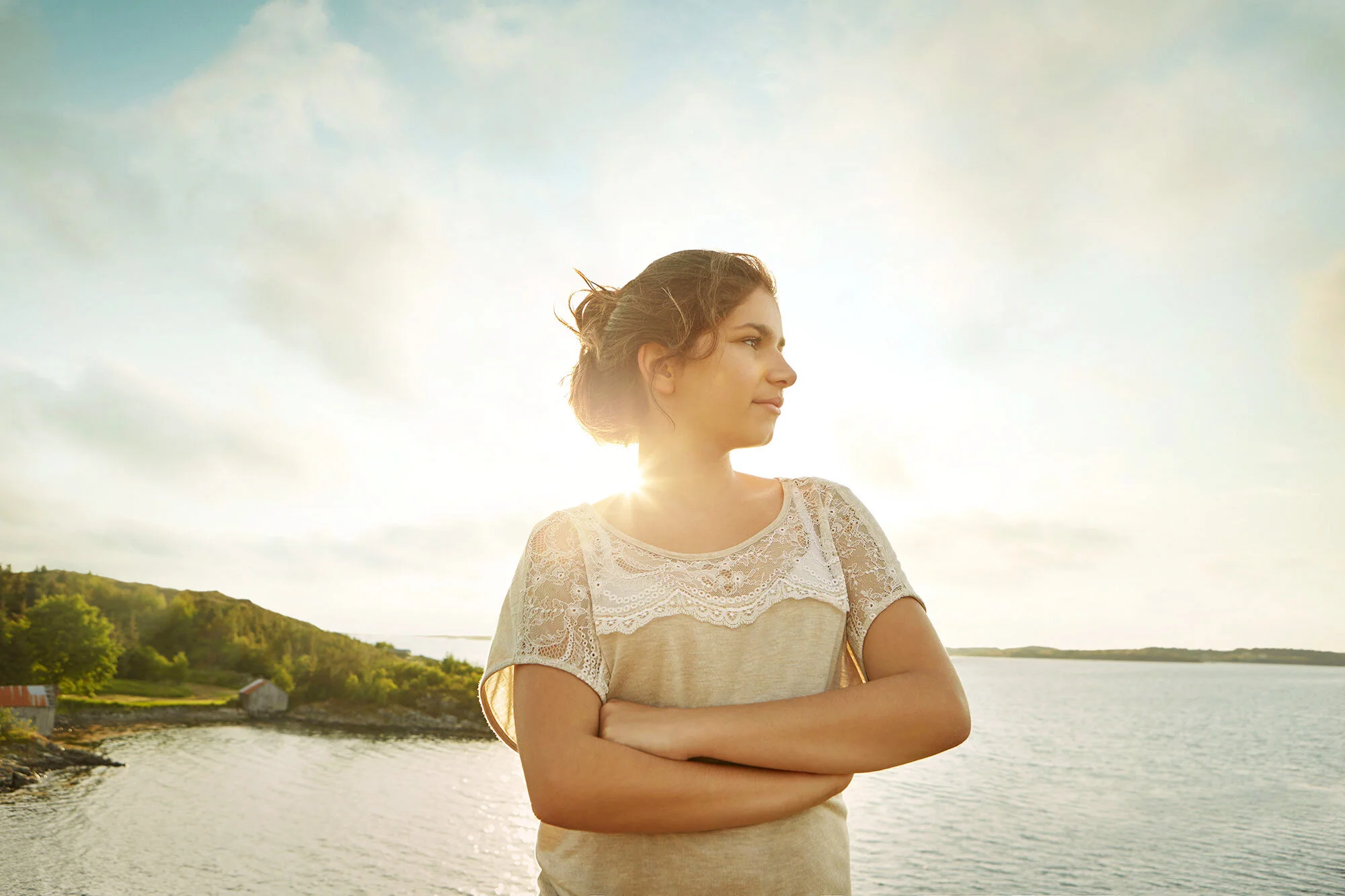 A young woman with long hair is standing outdoors near a body of water, crossing her arms and looking to the right, with the sun shining behind her and a landscape of trees and hills in the background at Hitra, Norway.