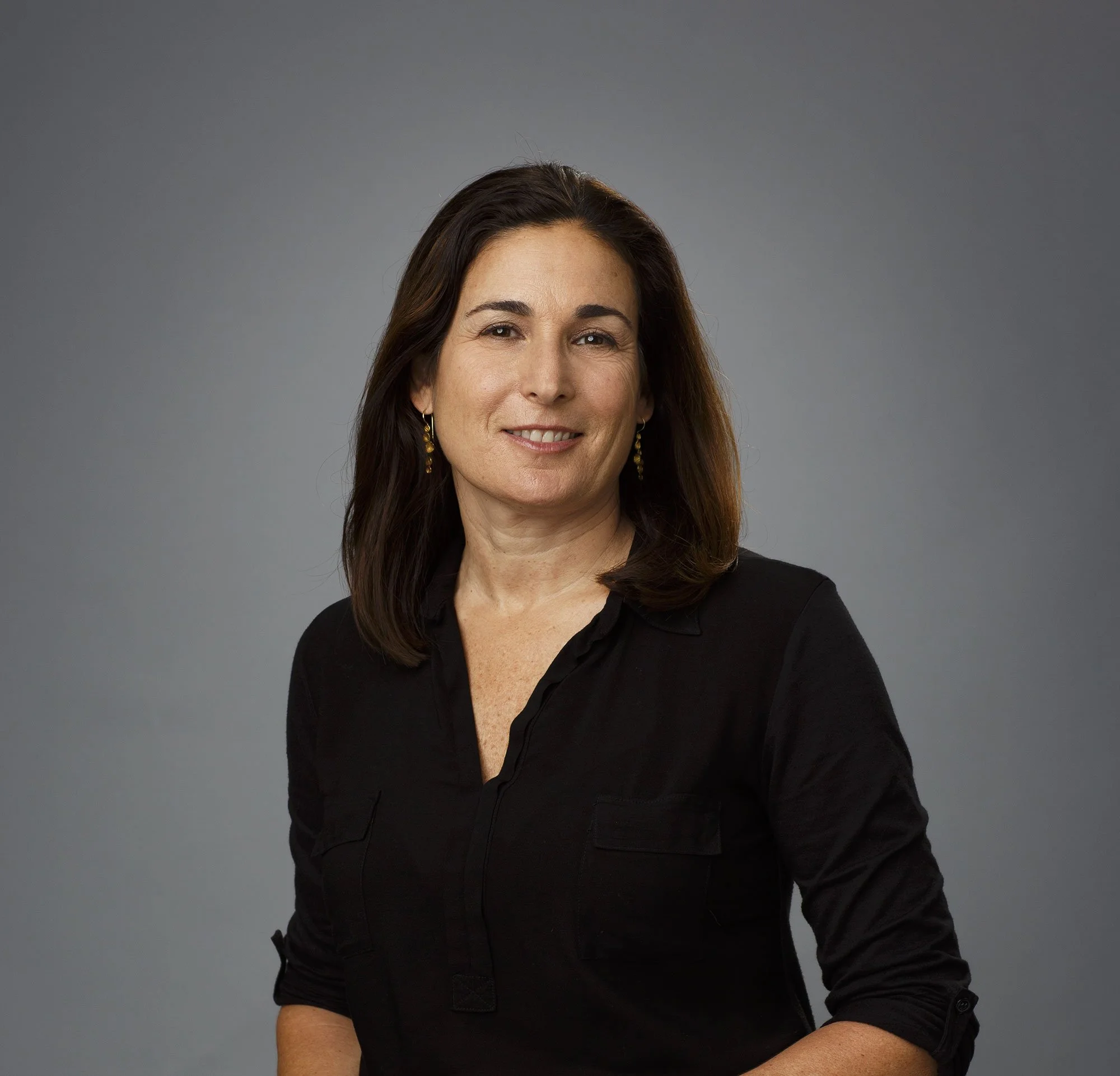 A white woman with shoulder-length dark hair wearing a black shirt and gold earrings, smiling slightly, against a plain gray background in San Francisco, CA.