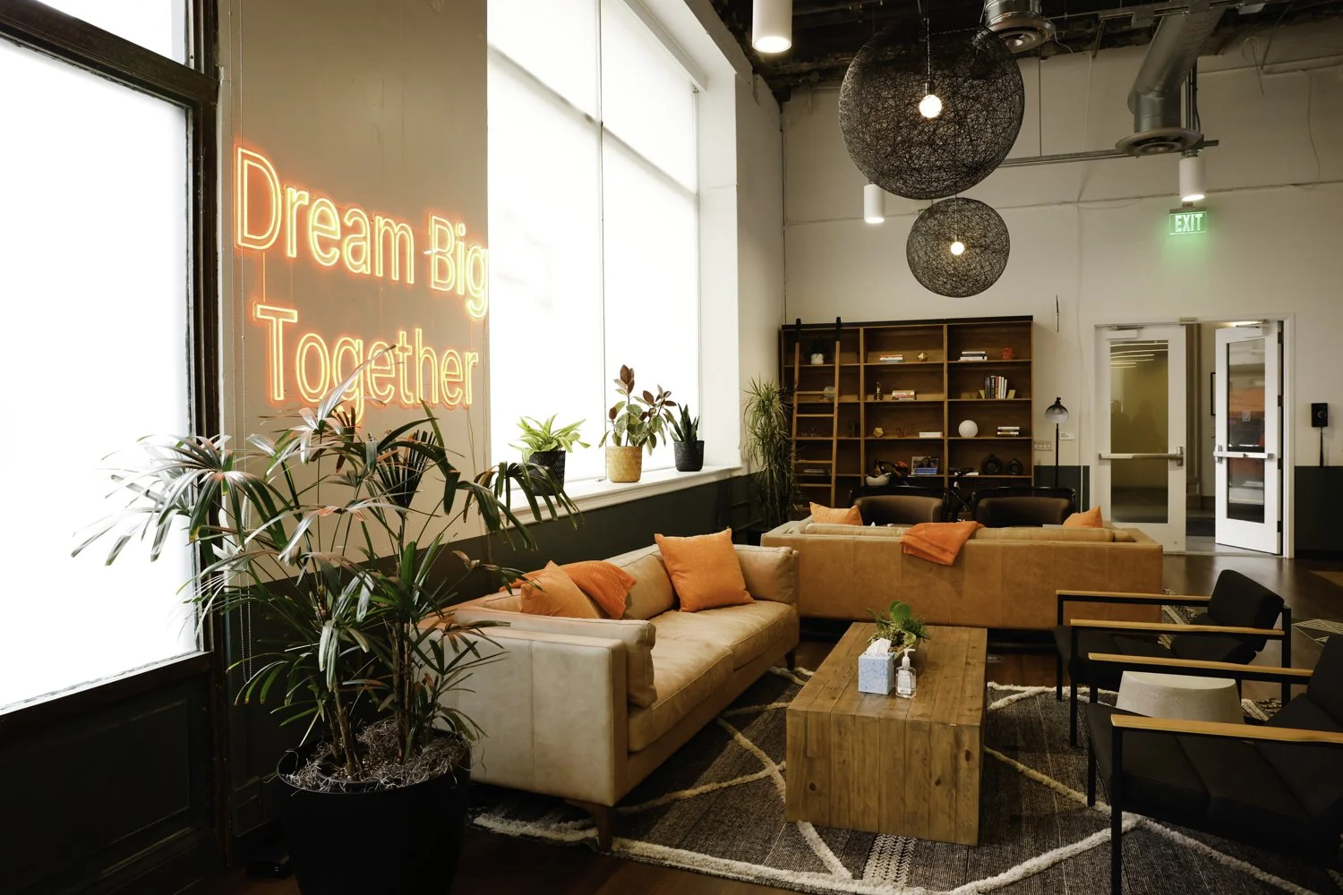 A modern lounge area with beige and black sofas, orange pillows, a large wooden coffee table, potted plants on the windowsill, a bookshelf filled with books and decor, black hanging spherical light fixtures, and a neon sign that reads 'Dream Big Toge
