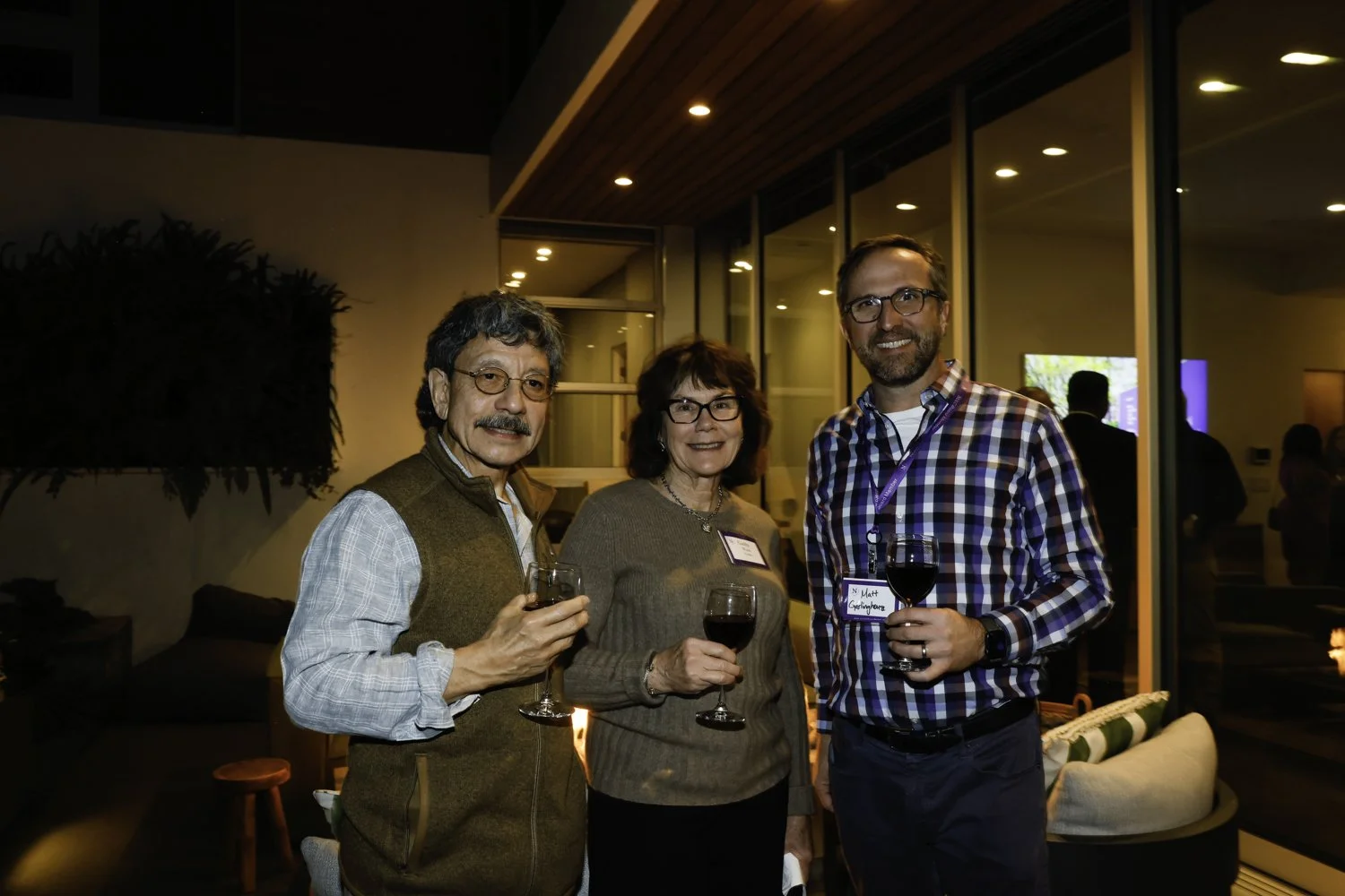 Three people, two men and one woman, smiling and holding glasses of red wine at a social gathering indoors, with a modern warm-lit background at Northwestern fundraiser in San Francisco, CA.