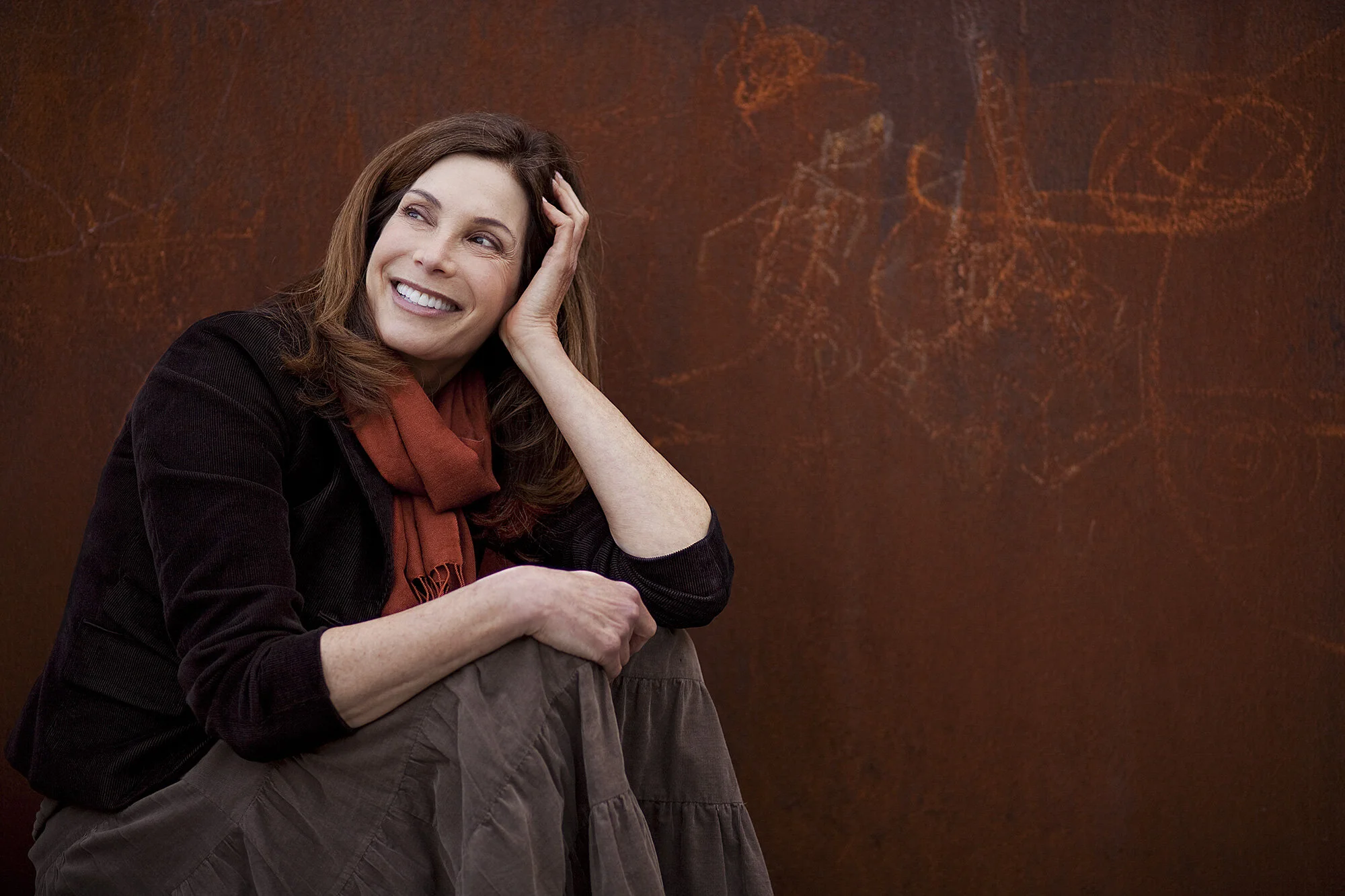 A smiling white middle-aged woman with brown hair, wearing a black jacket and an orange scarf, sitting against a dark reddish-brown textured background with abstract drawings in San Francisco, CA.