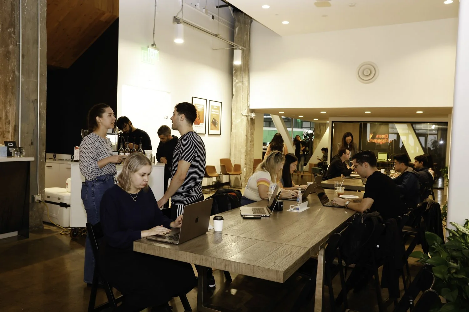 People working and socializing inside a modern coffee shop or co-working space, with some using laptops at a long table and others talking near the counter Brex headquarter in San Francisco, CA.