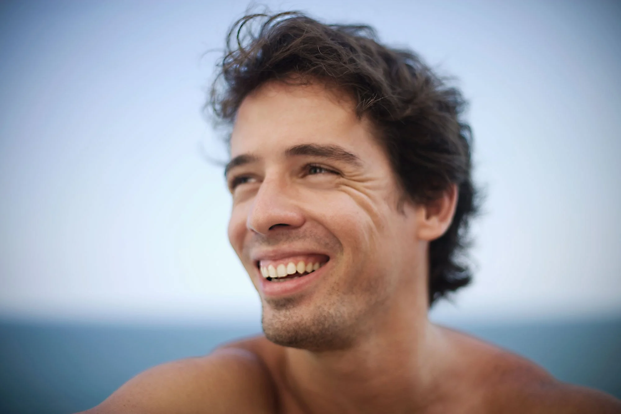Close-up of a smiling white Brazilian man with tousled dark hair, outdoors against a blurred sky background and the Atlantic ocean in at Ipanema beach in Rio de Janeiro, Brazil.