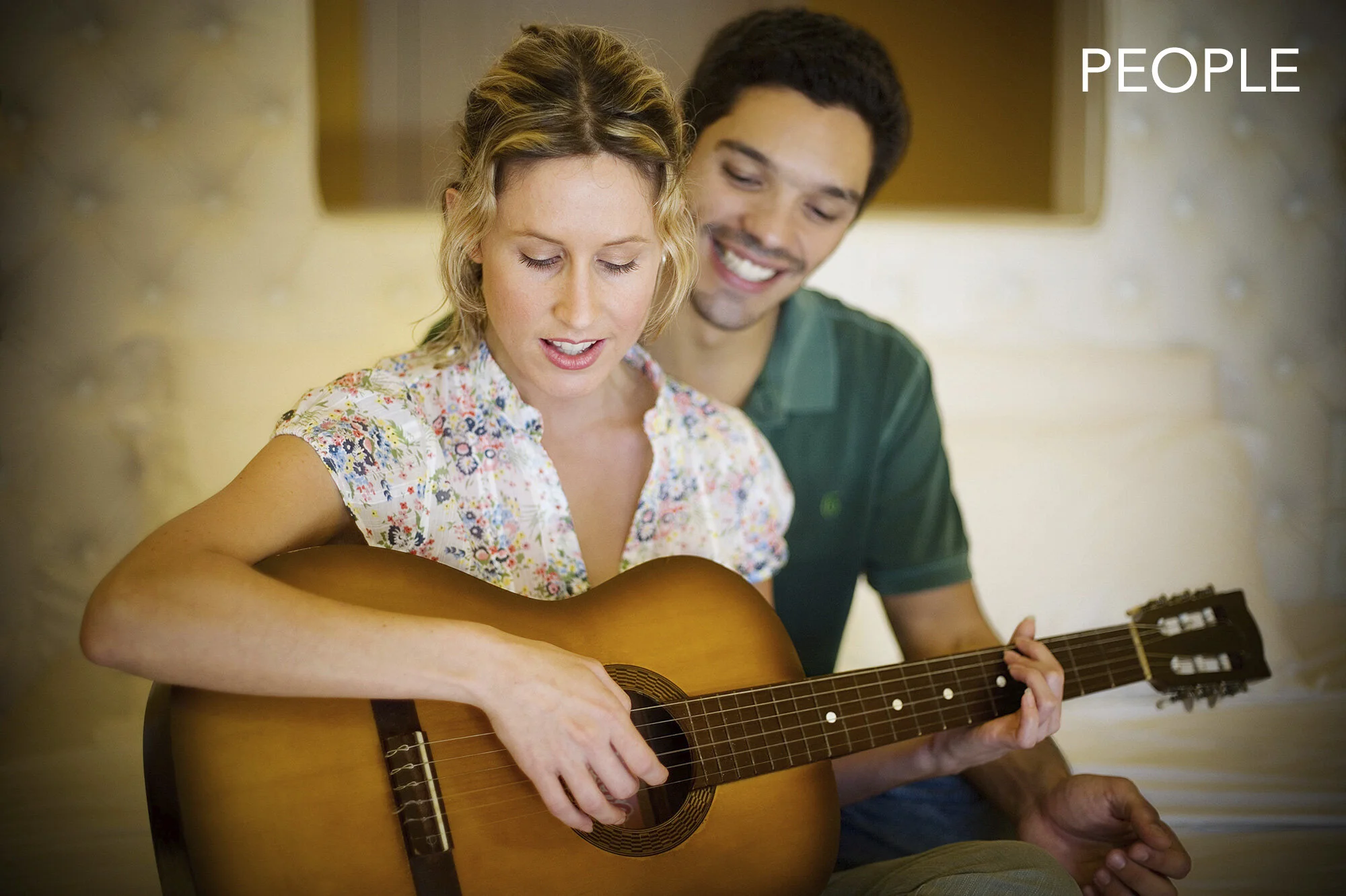 A woman playing an acoustic guitar with a man sitting behind her, smiling. They are indoors in hotel room, with soft lighting and neutral-colored background at La Suite boutique hotel in Rio de Janeiro, Brazil.