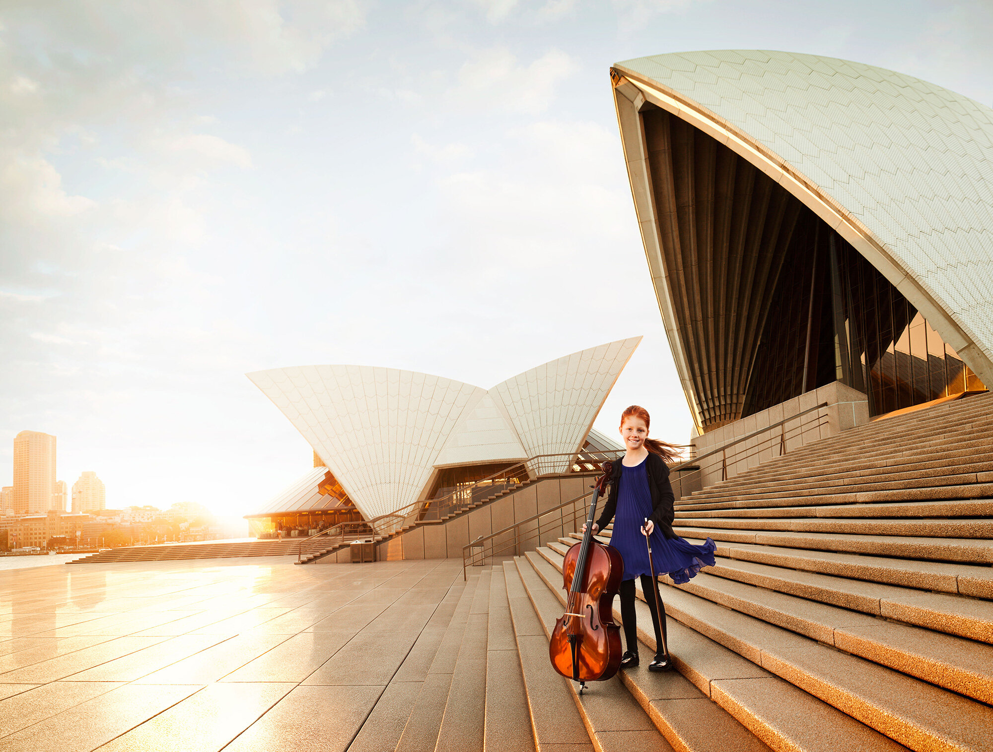 A red headed young girl in a blue dress and black jacket stands on stairs outside the Sydney Opera House, holding a cello and a bow, with the city skyline in the background at sunset in Sydney, Australia.