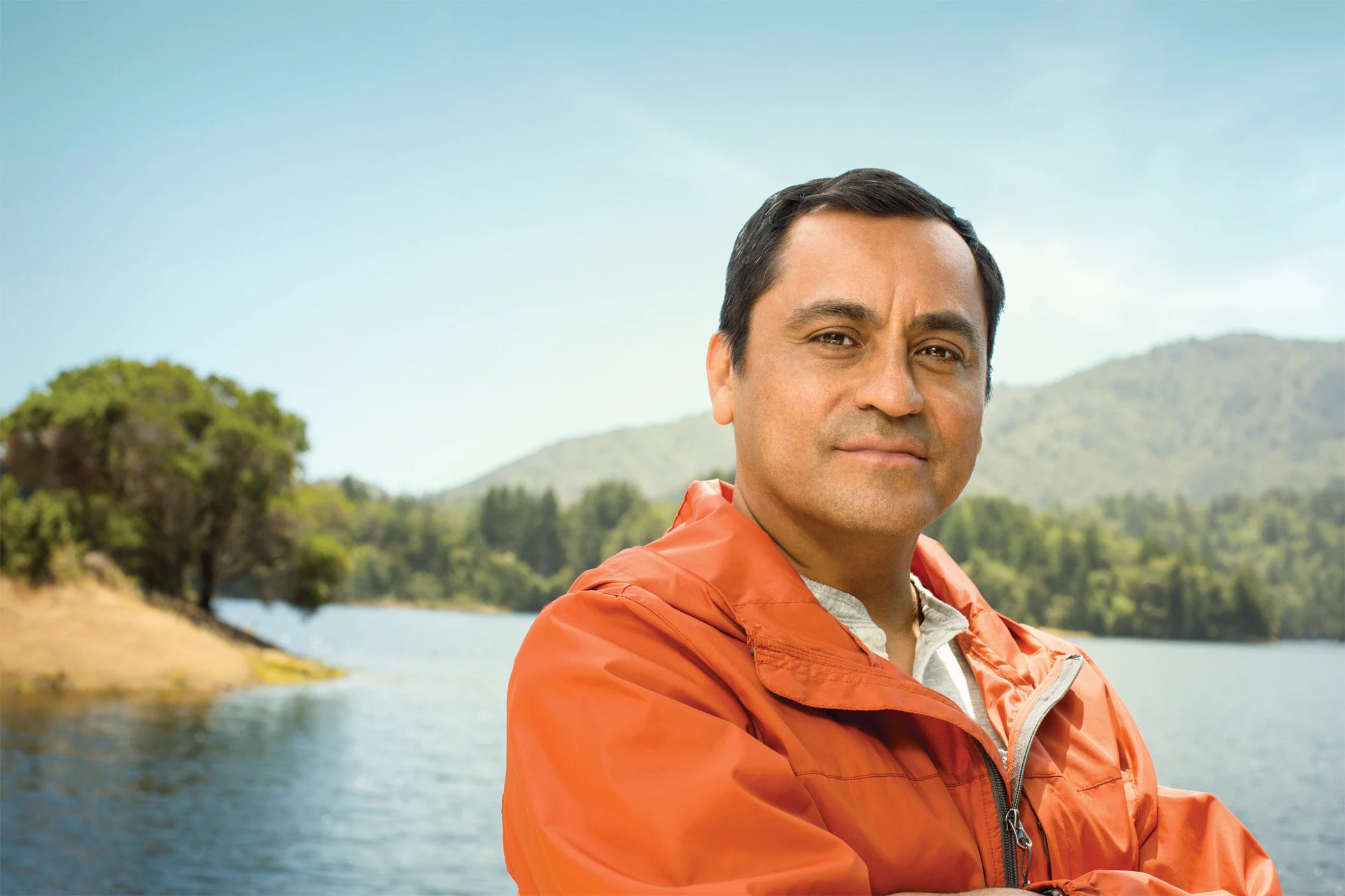 A Latino man in an orange jacket standing with his arms crossed  outdoors near a lake with trees and mountains in the background against a blue sky in San Francisco, CA.