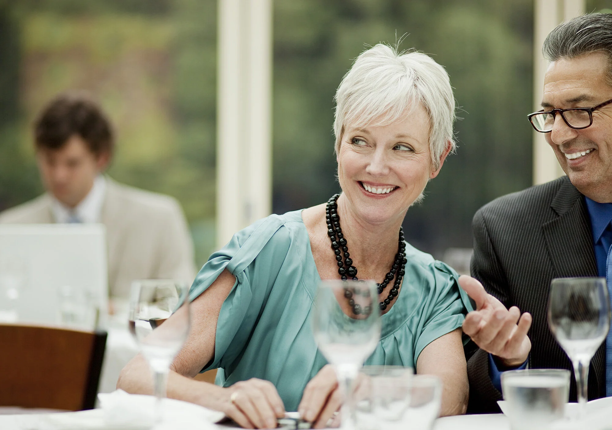 A smiling white middle-aged woman with short white hair wearing a turquoise dress and black beaded necklaces sitting at a table with empty wine glasses, with a man wearing glasses and a suit speaking to her. In the background, a person with curly hai