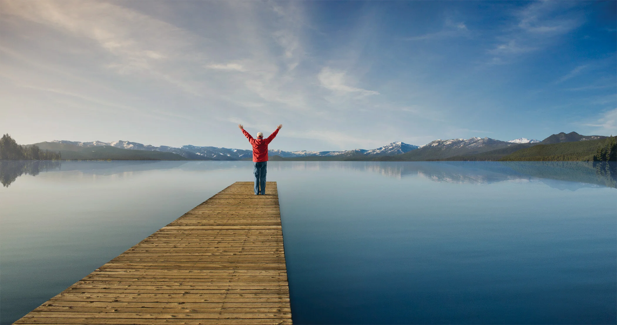 A man with red jacket and blue jeans on a wooden dock facing a calm lake with snow-capped mountains in the background, arms raised in the air at Lake Tahoe, CA.