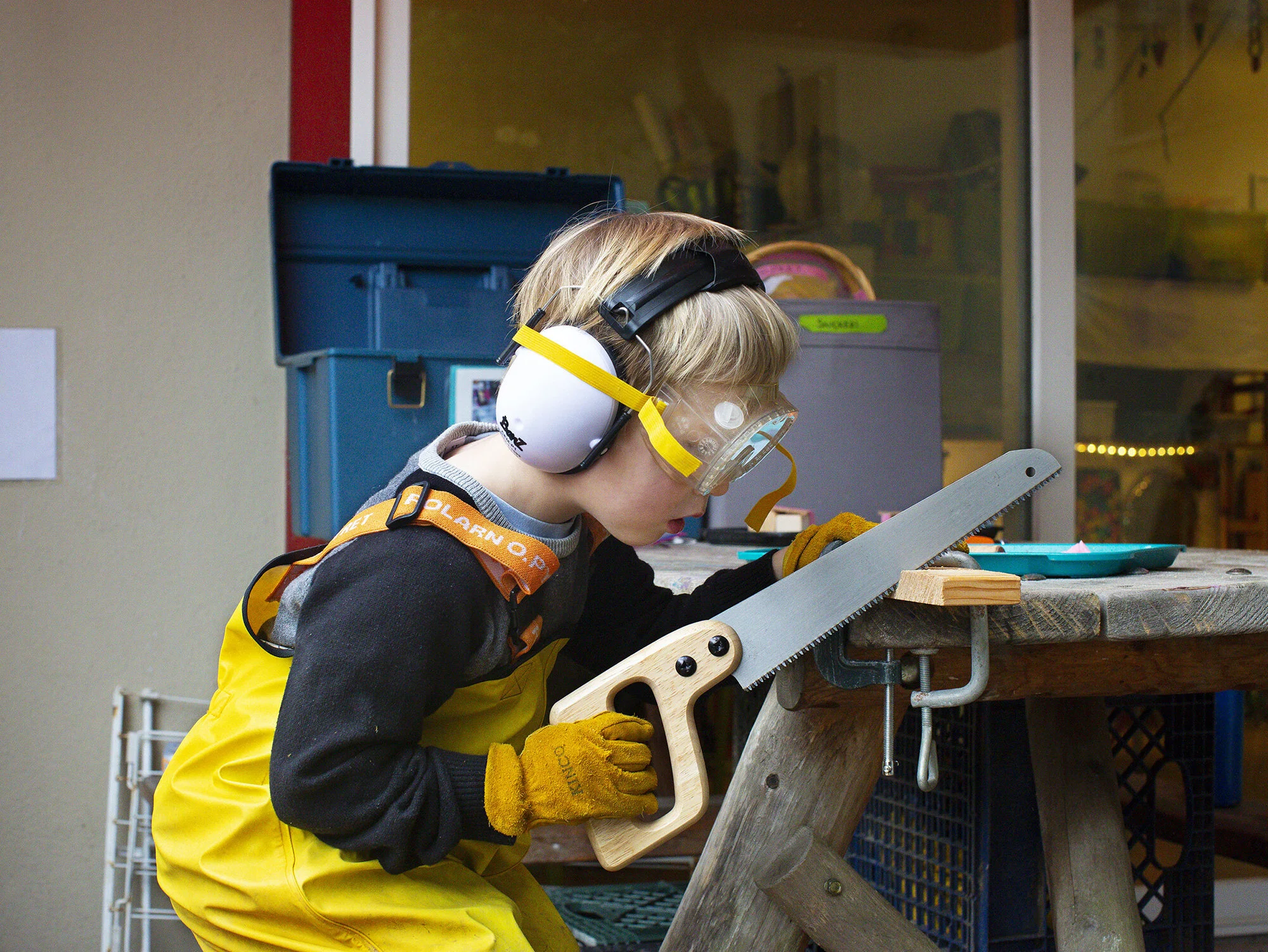 A young boy wearing ear protection, yellow rain pants, safety goggles, gloves, and a yellow apron is using a hand saw to cut a piece of wood on a workbench at the The Scandinavian School in San Francisco, CA.