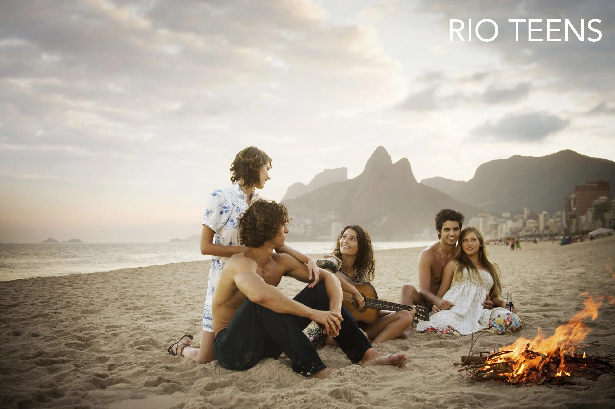 Group of young people sitting on the beach near a small bonfire at Ipanema beach in Rio de Janeiro, Brazil, with mountains and city buildings in the background at sunset, one holding a guitar, others smiling and talking.