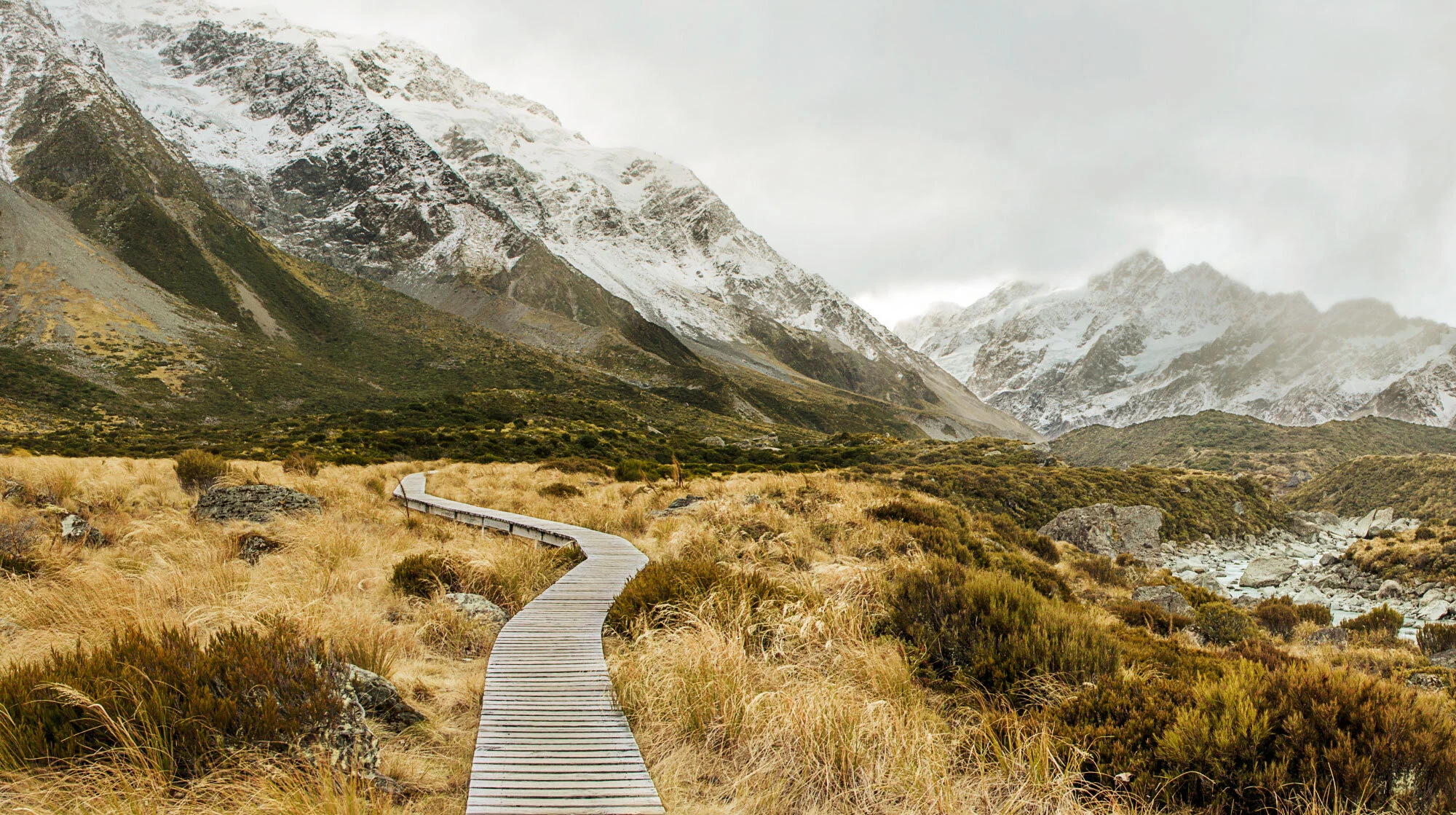 A winding wooden pathway through a grassy mountain landscape with snow-capped peaks in the background in New Zealand.