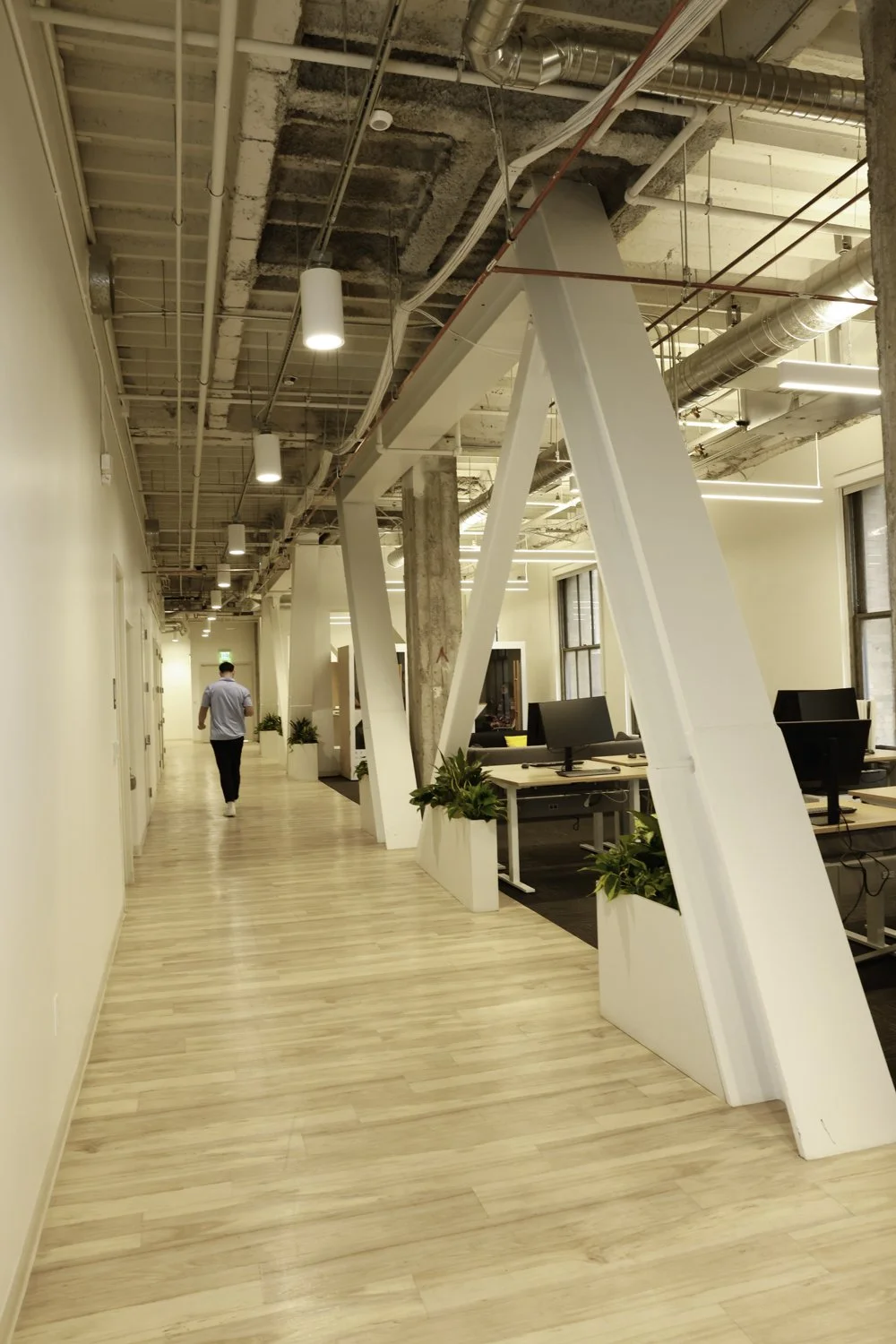 Modern office with wooden flooring, exposed ceiling pipes, large windows, desks with computers, and potted plants Brex headquarter in San Francisco, CA.